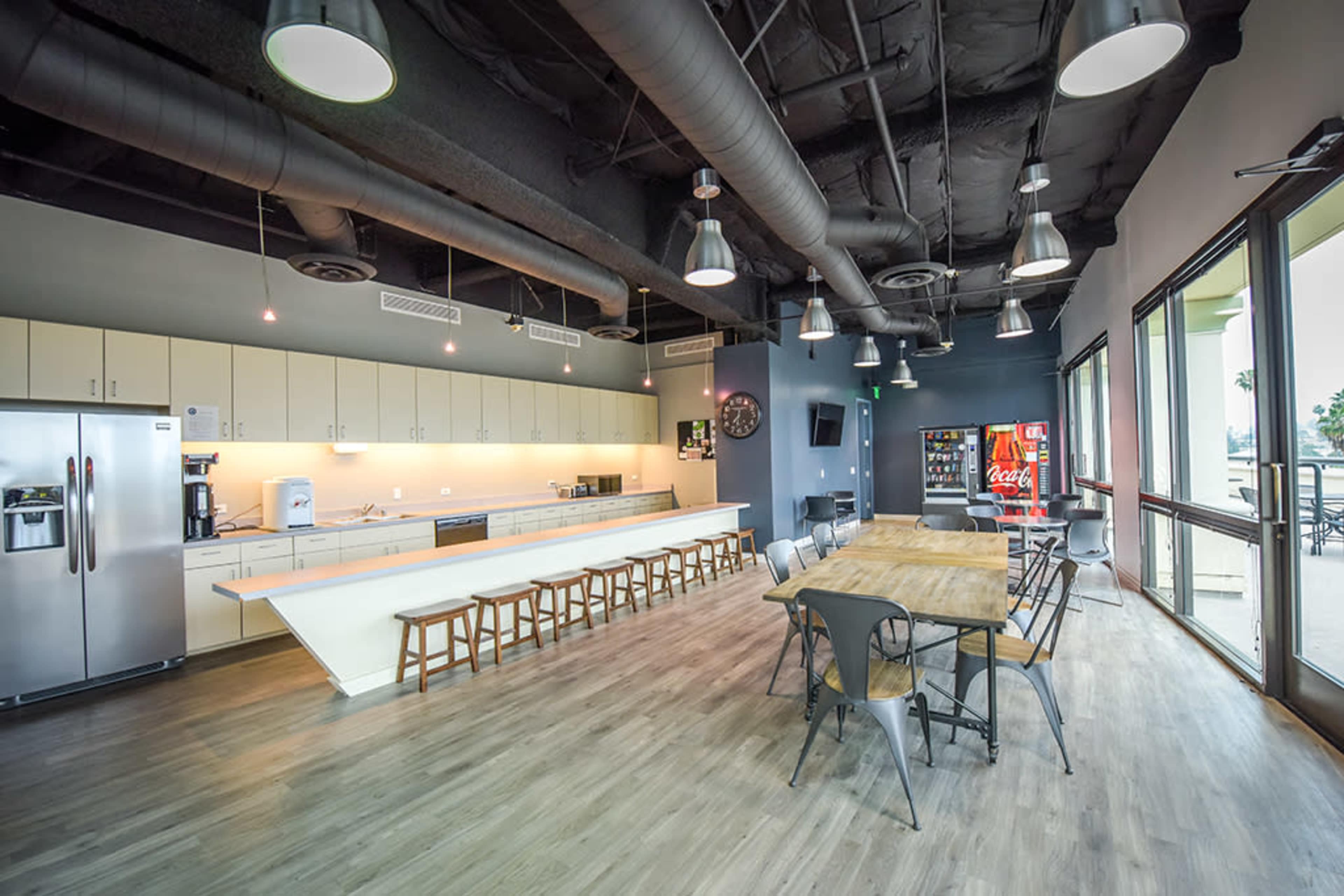 A modern kitchen and dining area with a long countertop, metal chairs, a wooden table, and a vending machine.