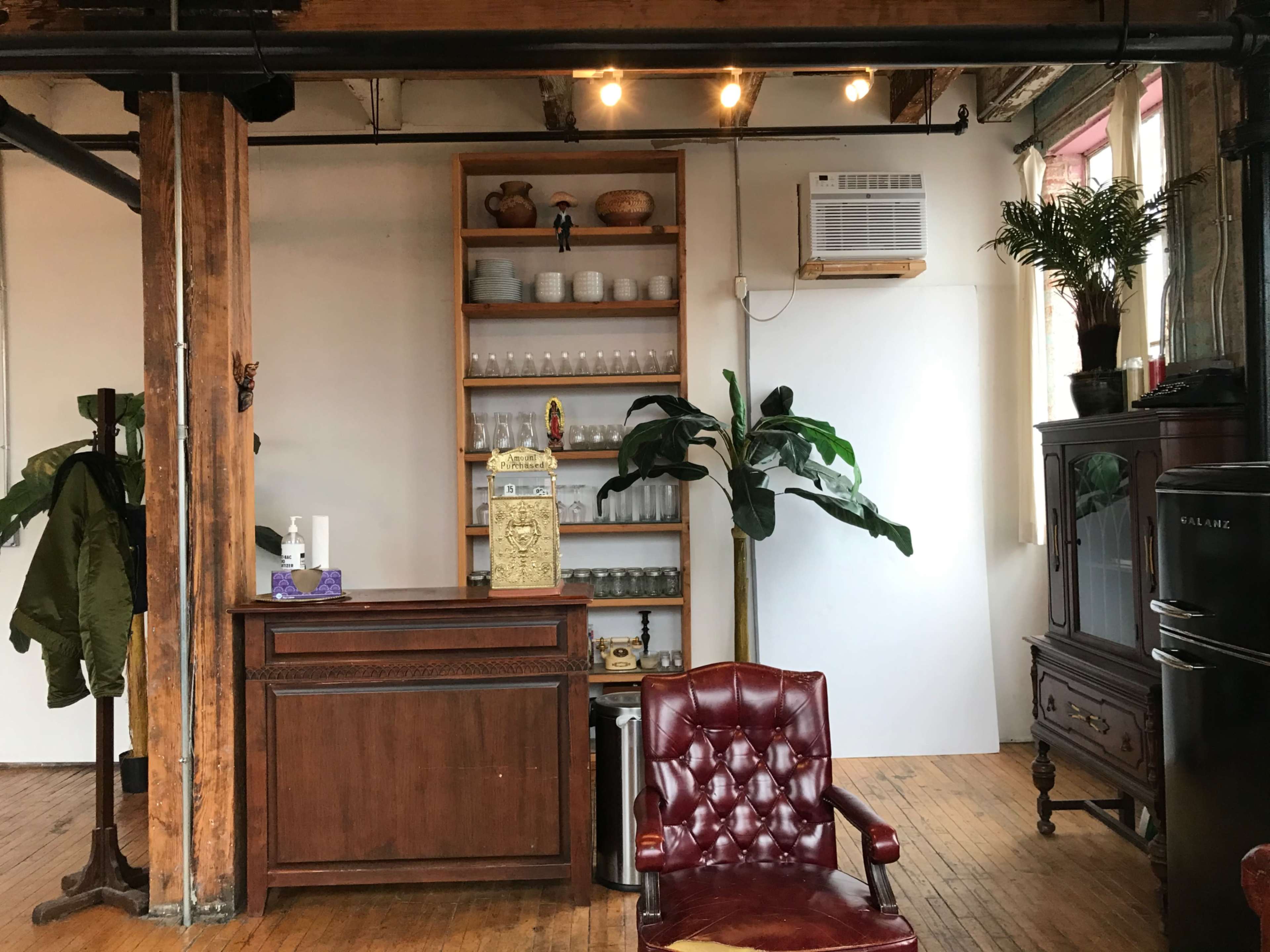 A room with wooden floors, a vintage brown chair, a wooden bar-like section with shelves of glassware, and decorative plants near a white wall and an air conditioning unit.