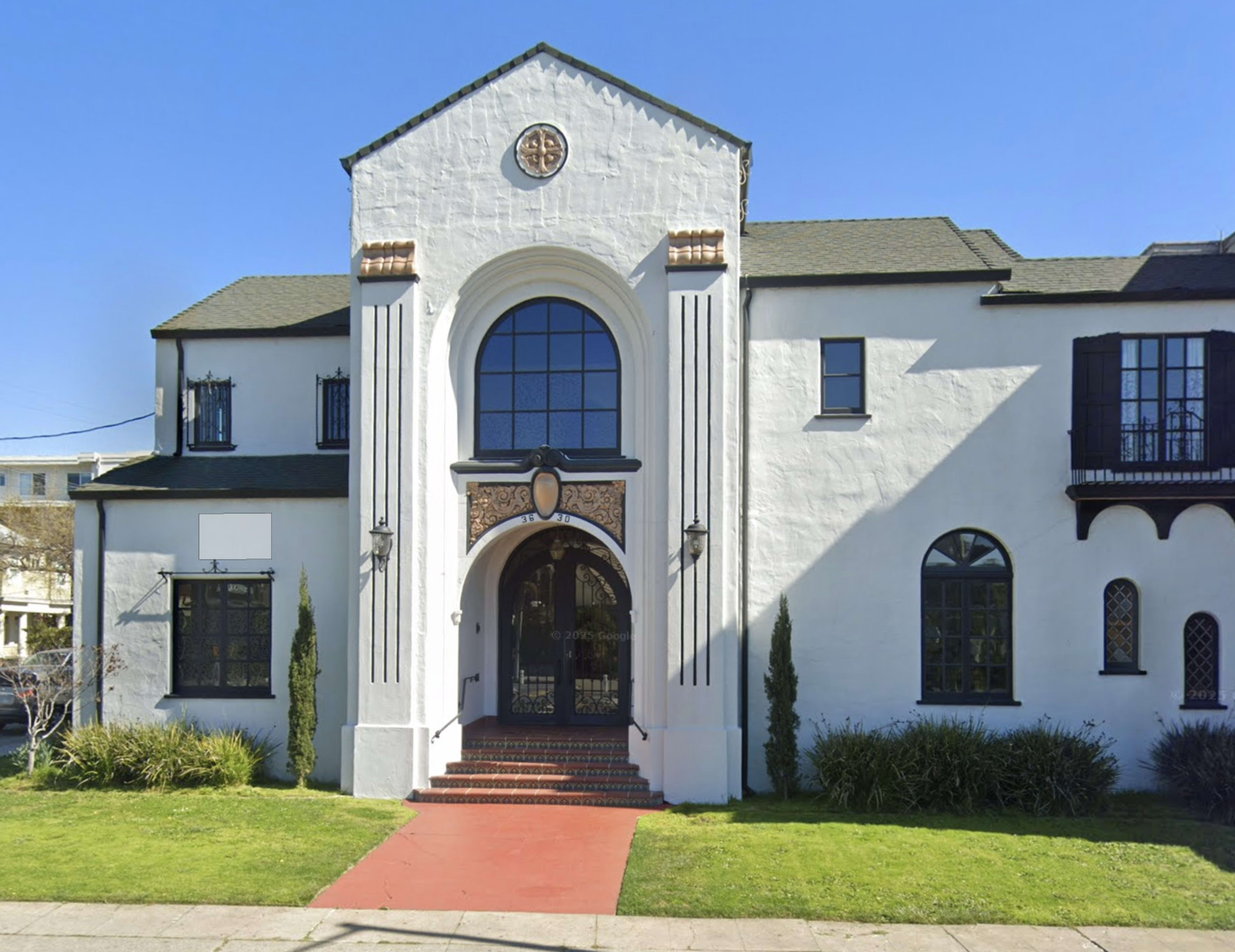 The image shows a large, two-story white house with decorative features, a prominent arched entrance, and a red pathway leading to the front door.