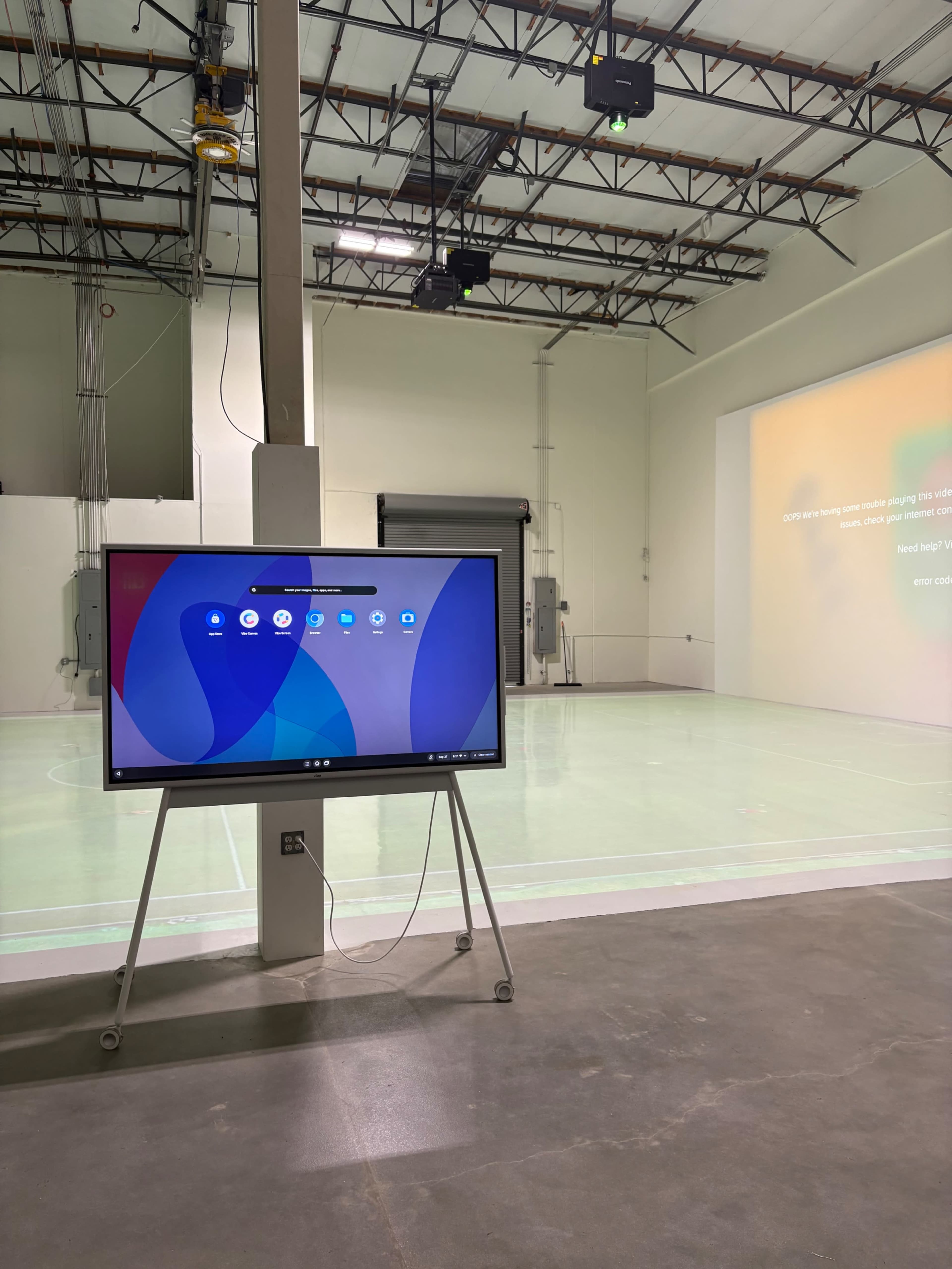 The image shows a large display screen mounted on a stand, positioned in a spacious, empty room with green-tinted flooring and bright overhead lights.