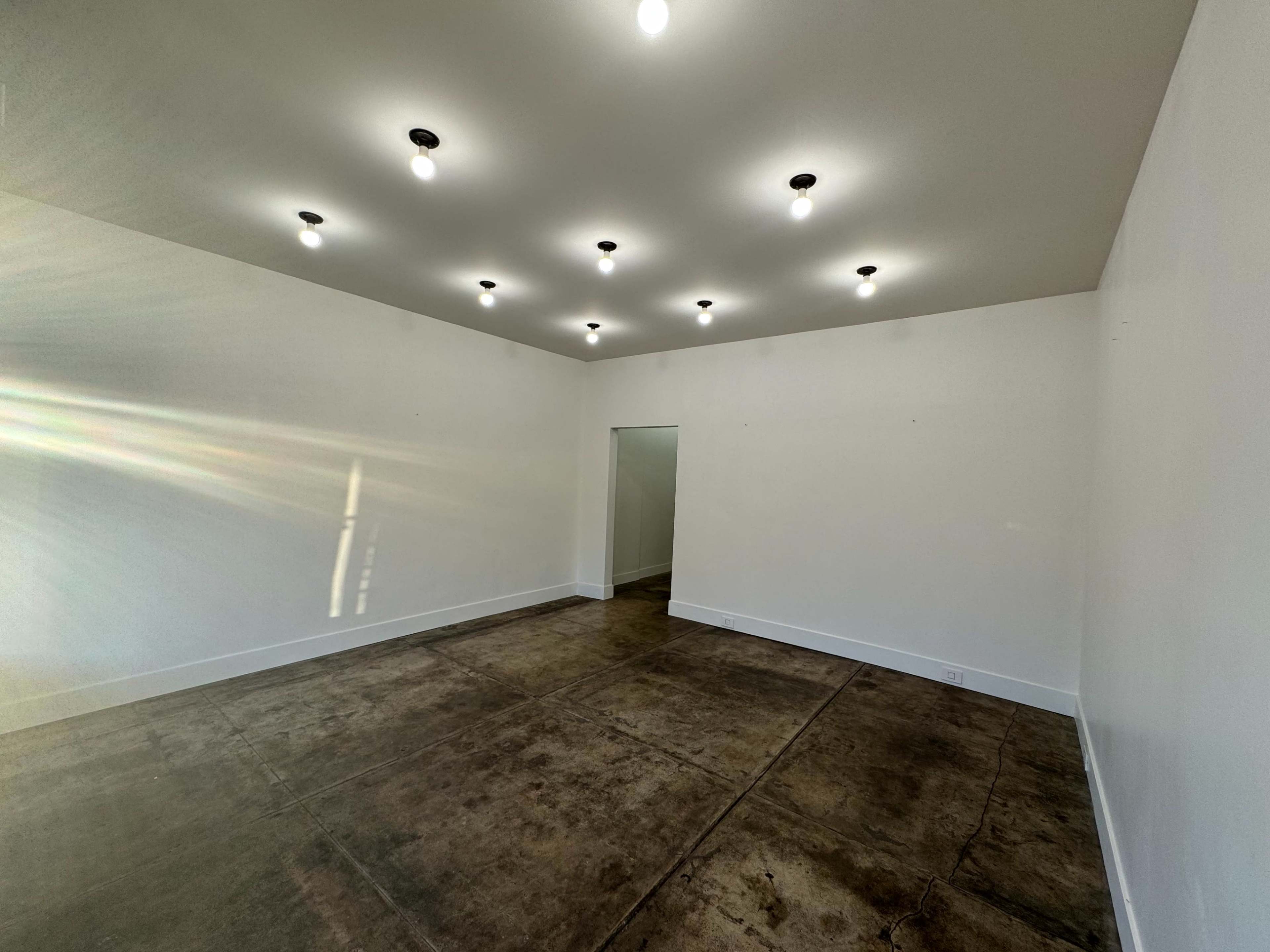 The image shows a large, empty room with a concrete floor, white walls, and several ceiling lights.