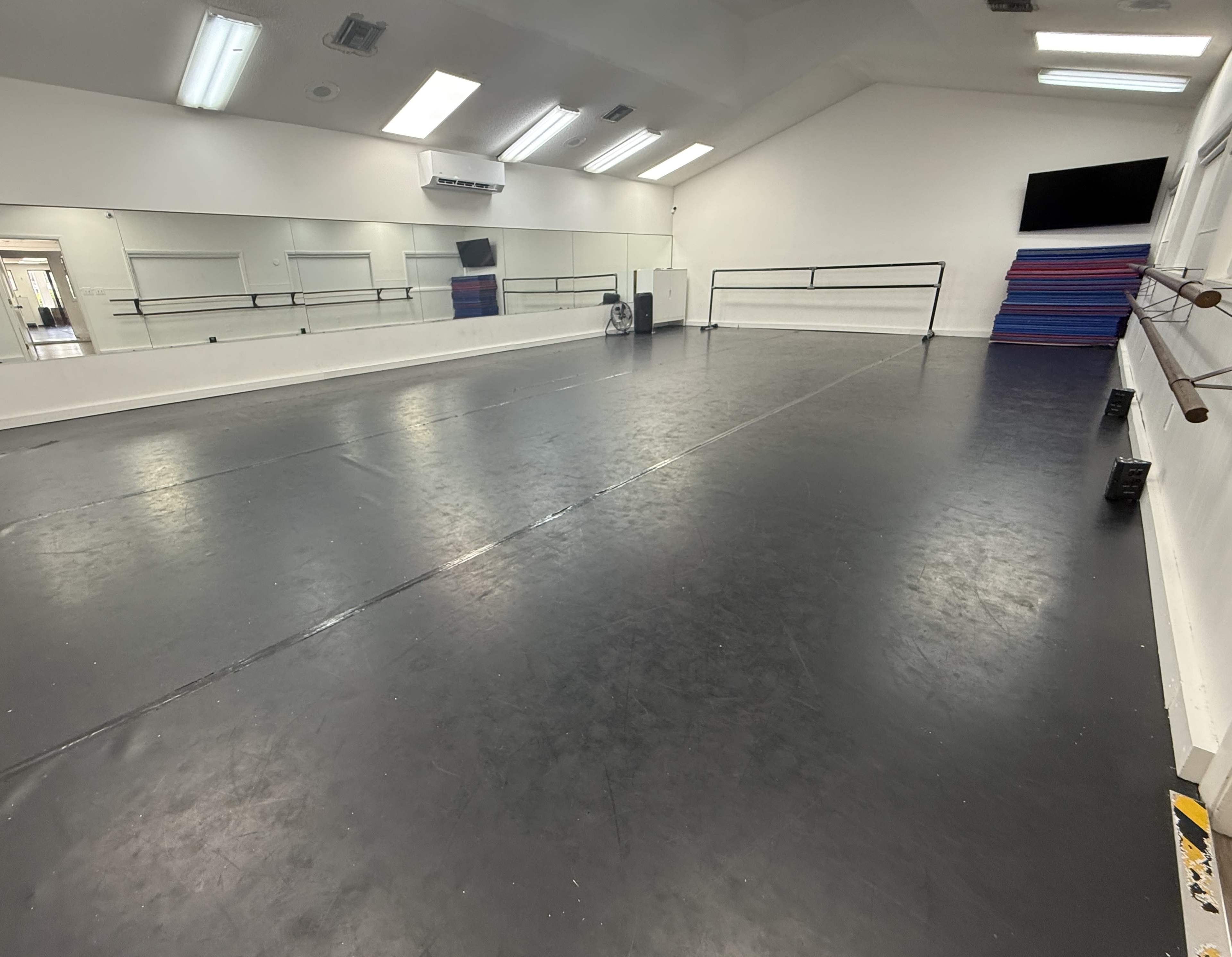 The image shows an empty dance studio with a mirrored wall, ballet bars, and a television mounted on the wall.
