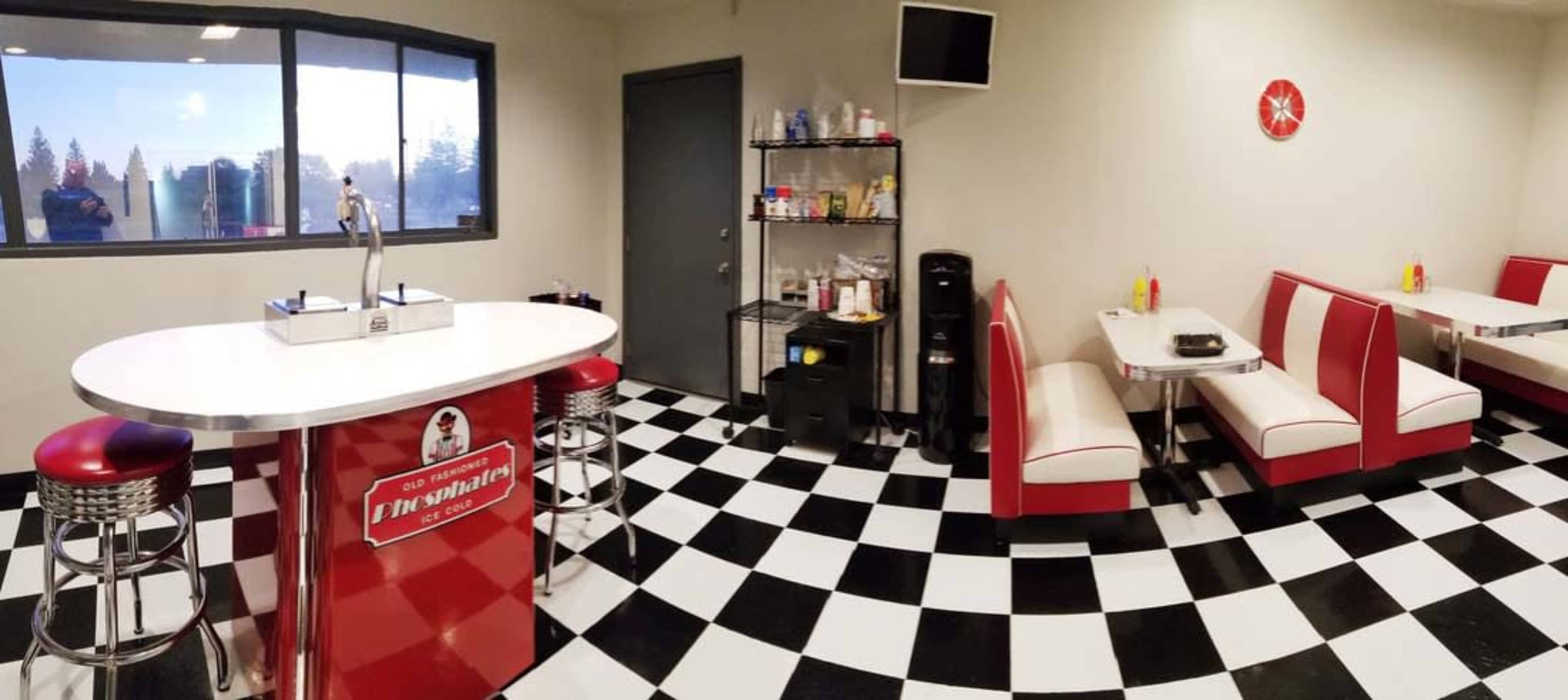 The image shows a retro-style diner featuring checkered floors, red and white seating booths, a bar with stools, and a counter with a beverage tap.
