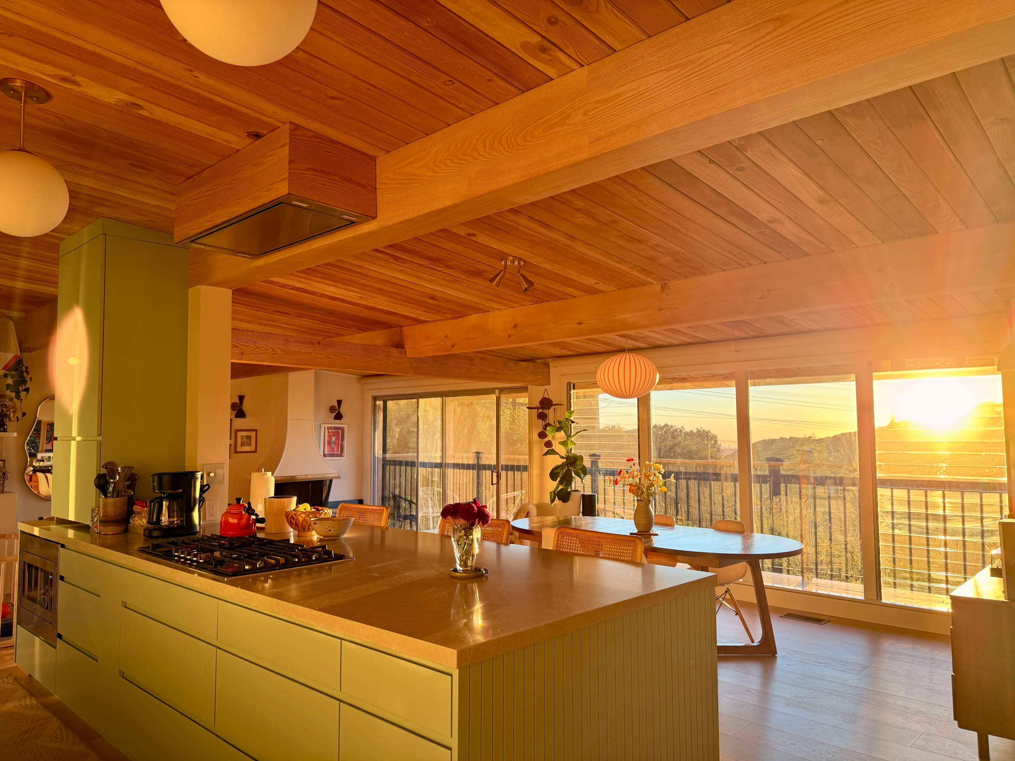A modern kitchen with a wooden ceiling features a green island, large windows allowing sunlight to pour in, and a dining table with a scenic view of the outdoors.