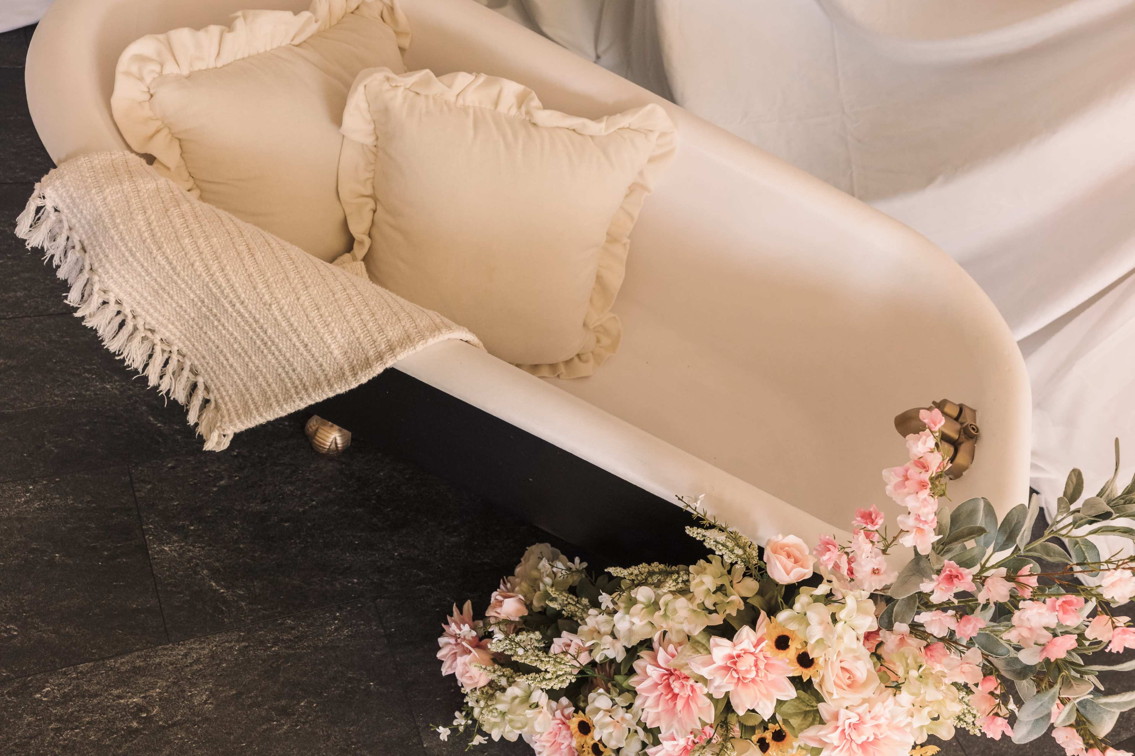 A vintage freestanding bathtub with two decorative pillows and a textured blanket, accompanied by a bouquet of pink and white flowers.