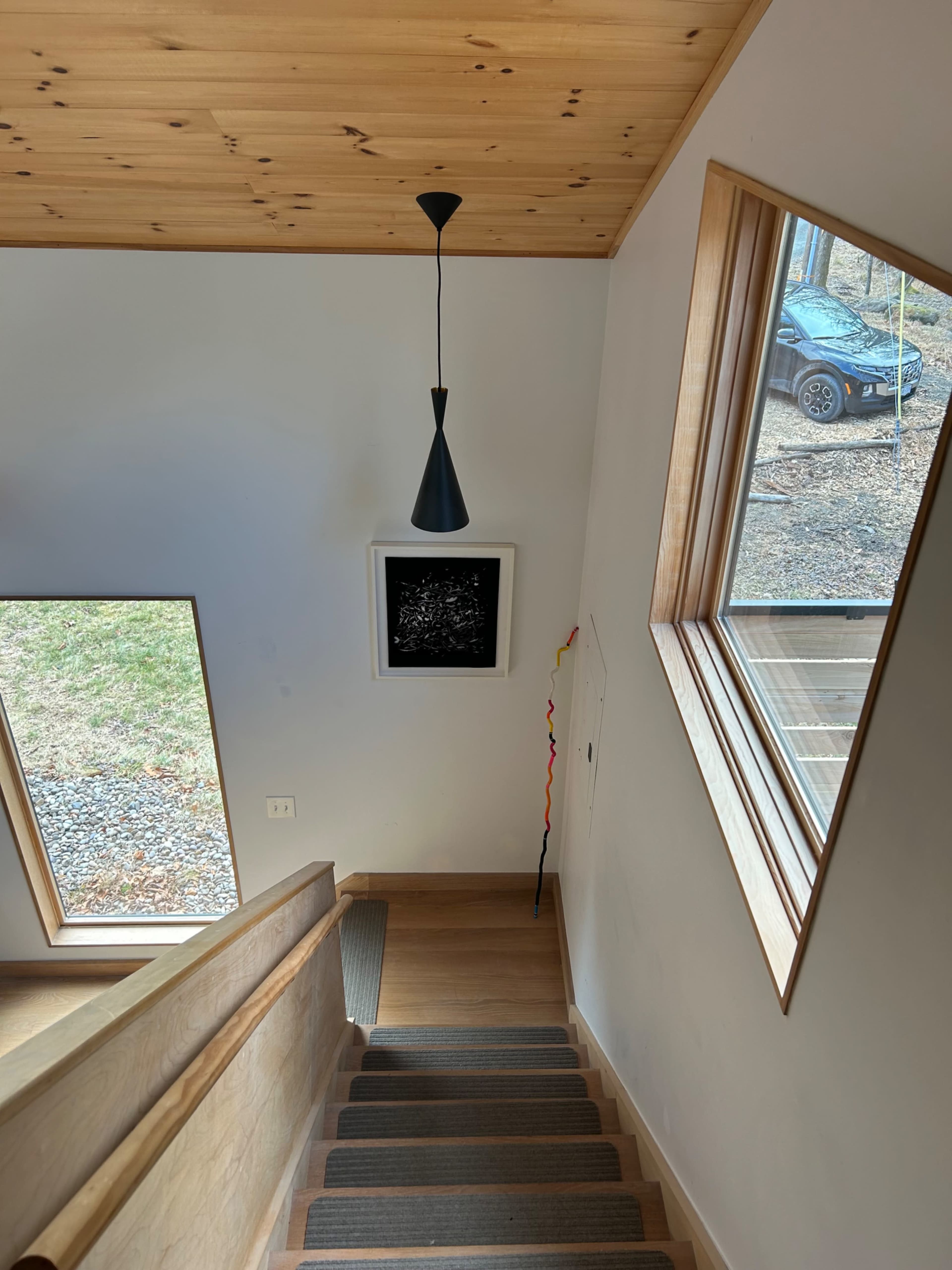 A staircase leads down from a wooden ceiling, with a pendant light hanging above and a black-and-white artwork on the wall, alongside large windows revealing a car parked outside.