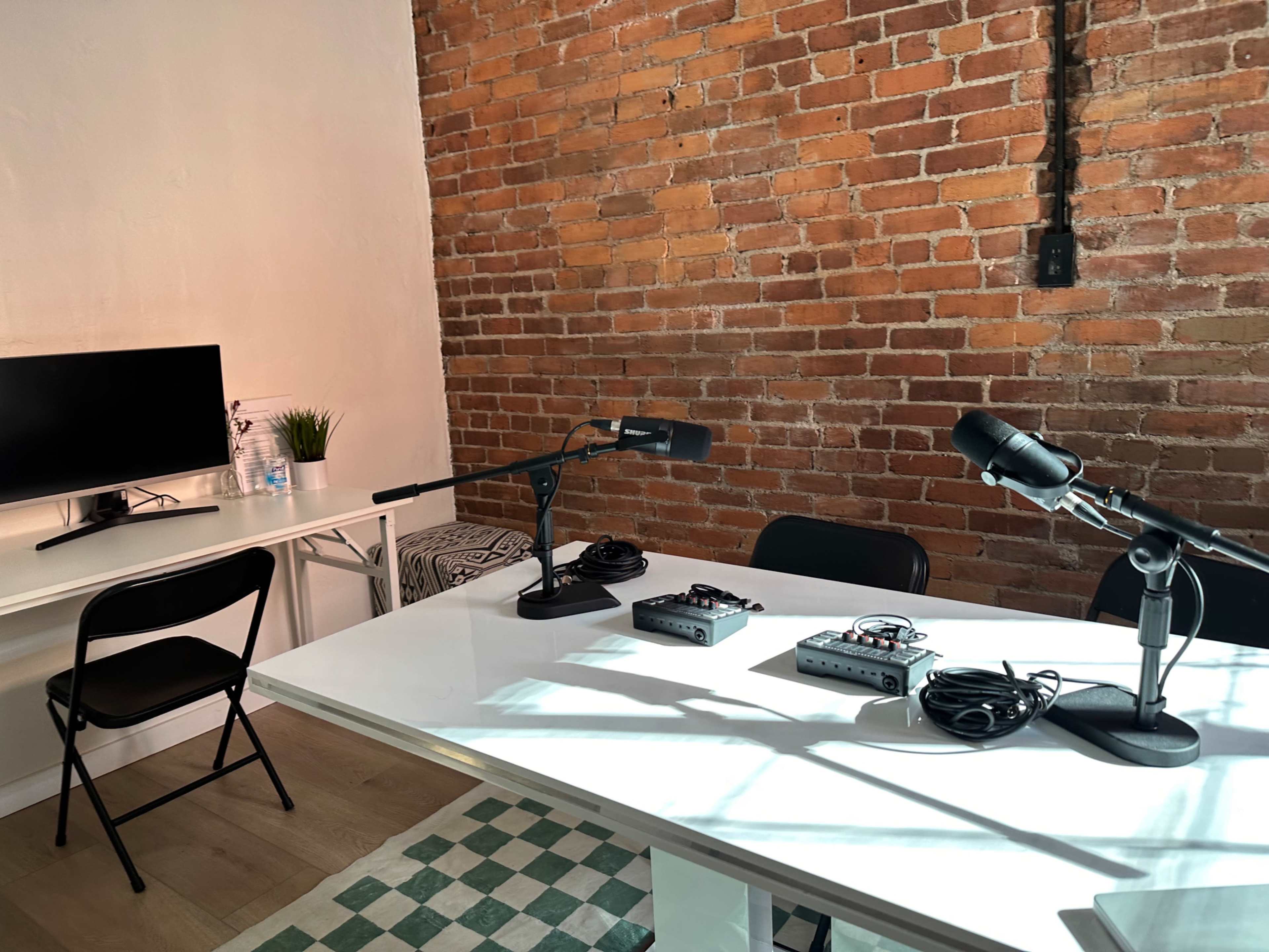 A minimalist podcast recording studio featuring a white table with three microphones and audio interfaces, against a backdrop of exposed brick walls and a small television on the side.
