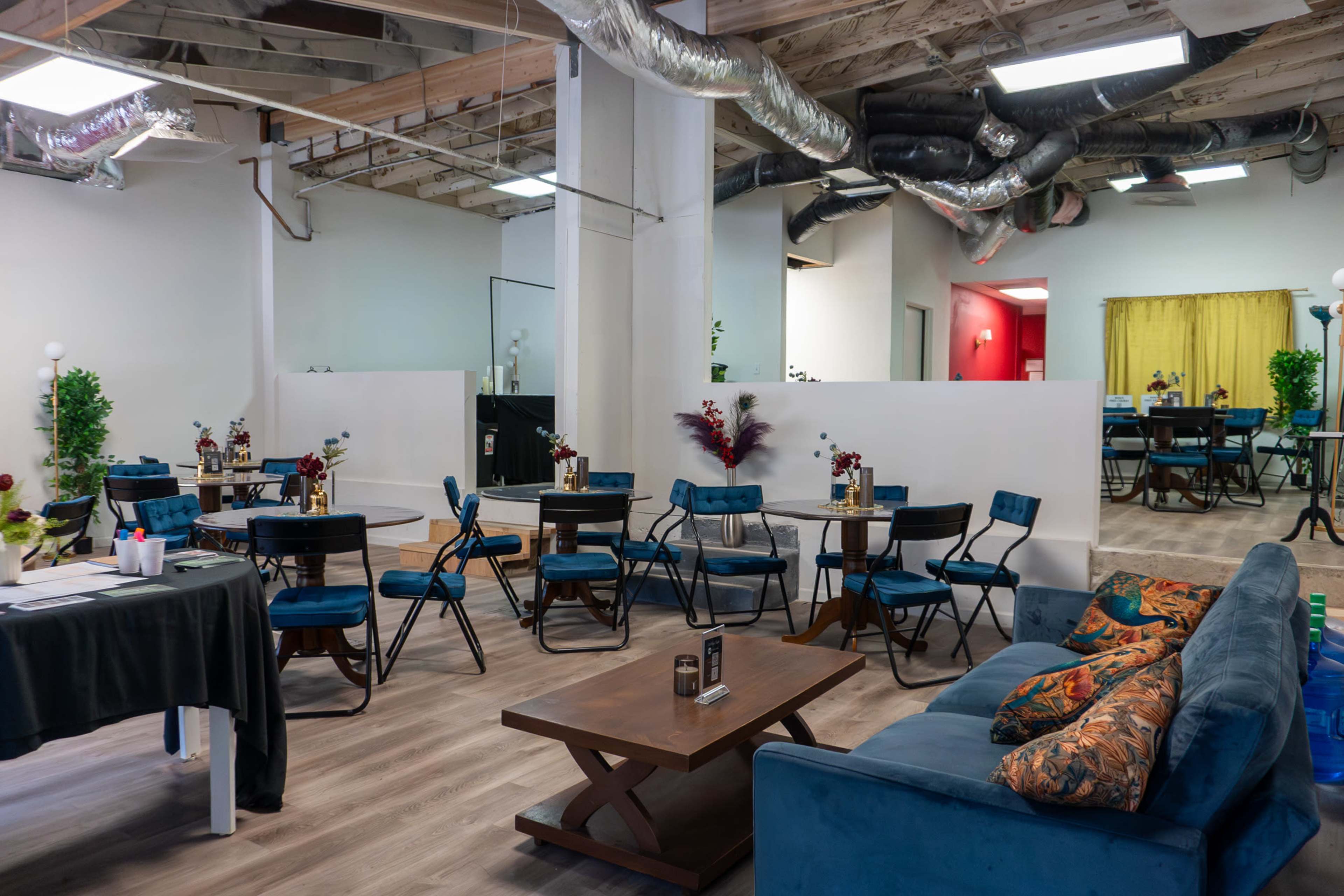 The image shows an office or communal space with several tables and chairs, a blue couch, and plants, featuring modern decor and distinct color themes in different sections.