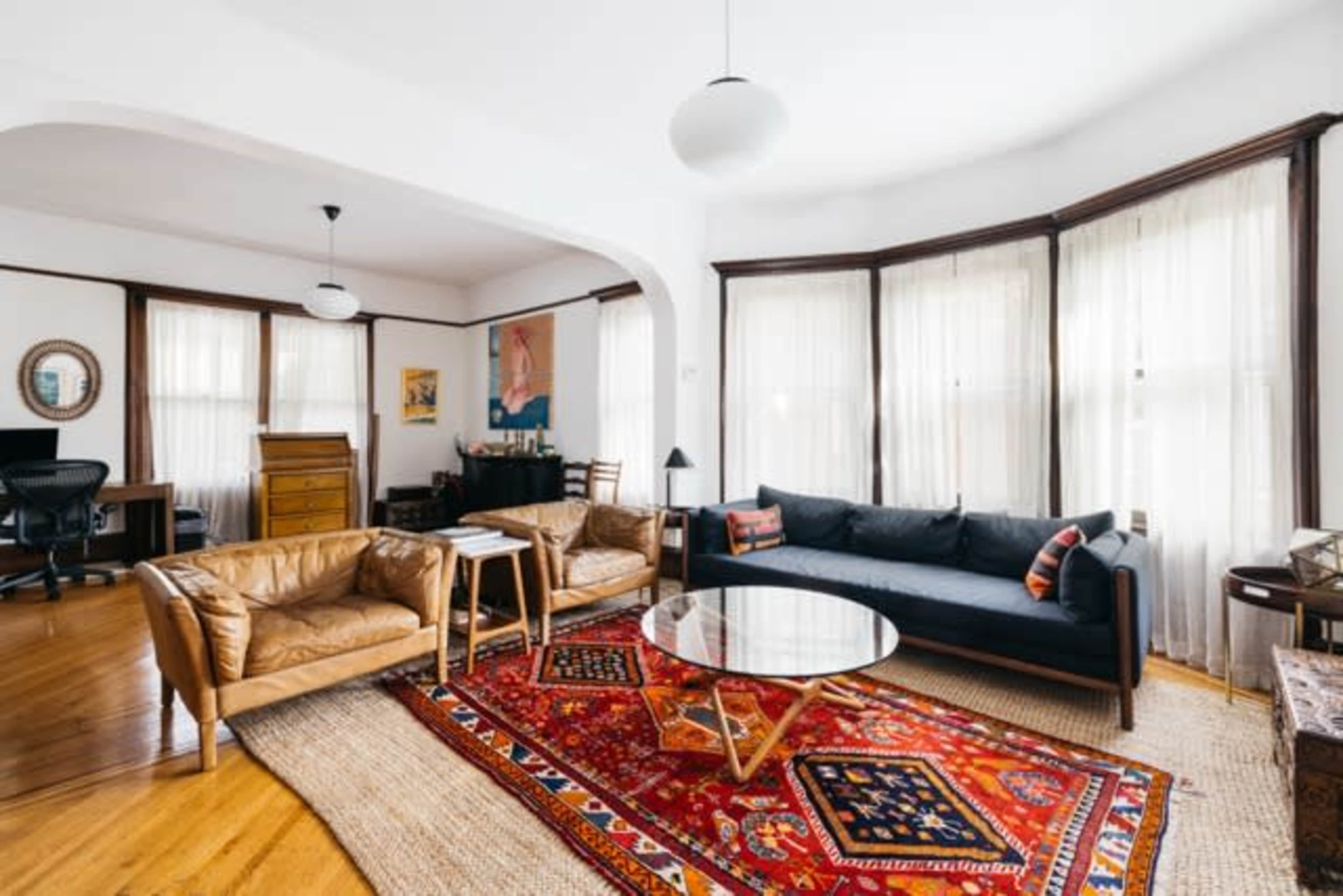 The image shows a bright living room featuring two tan leather armchairs, a dark blue sofa, a round coffee table on a colorful area rug, and large windows with sheer curtains.