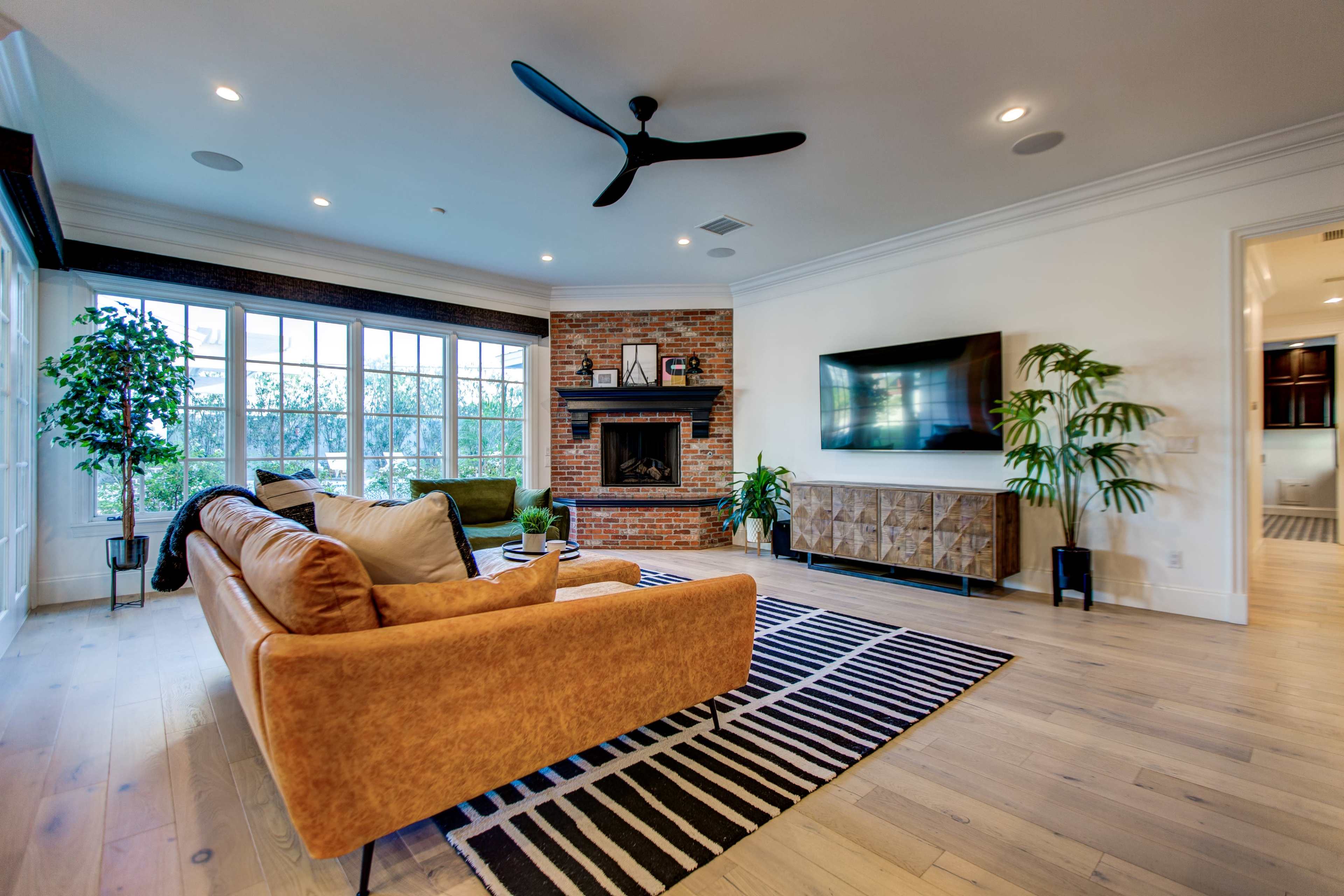 A modern living room features a brown sofa facing a fireplace and a mounted television, with large windows allowing natural light to fill the space.