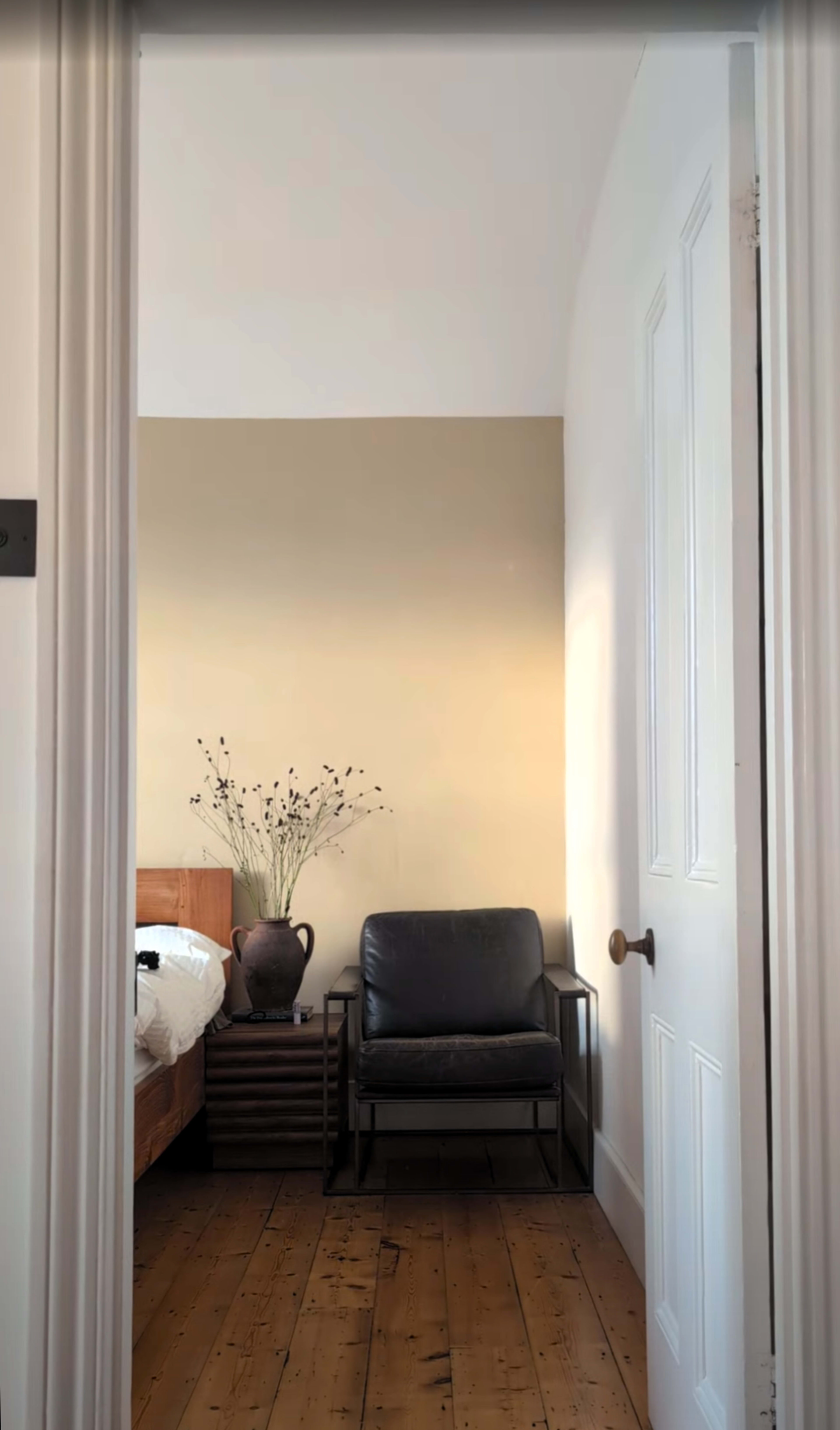 The image shows a view through an open door into a room with a bed, a chair, a wooden storage unit, and a vase with dried flowers on a surface.
