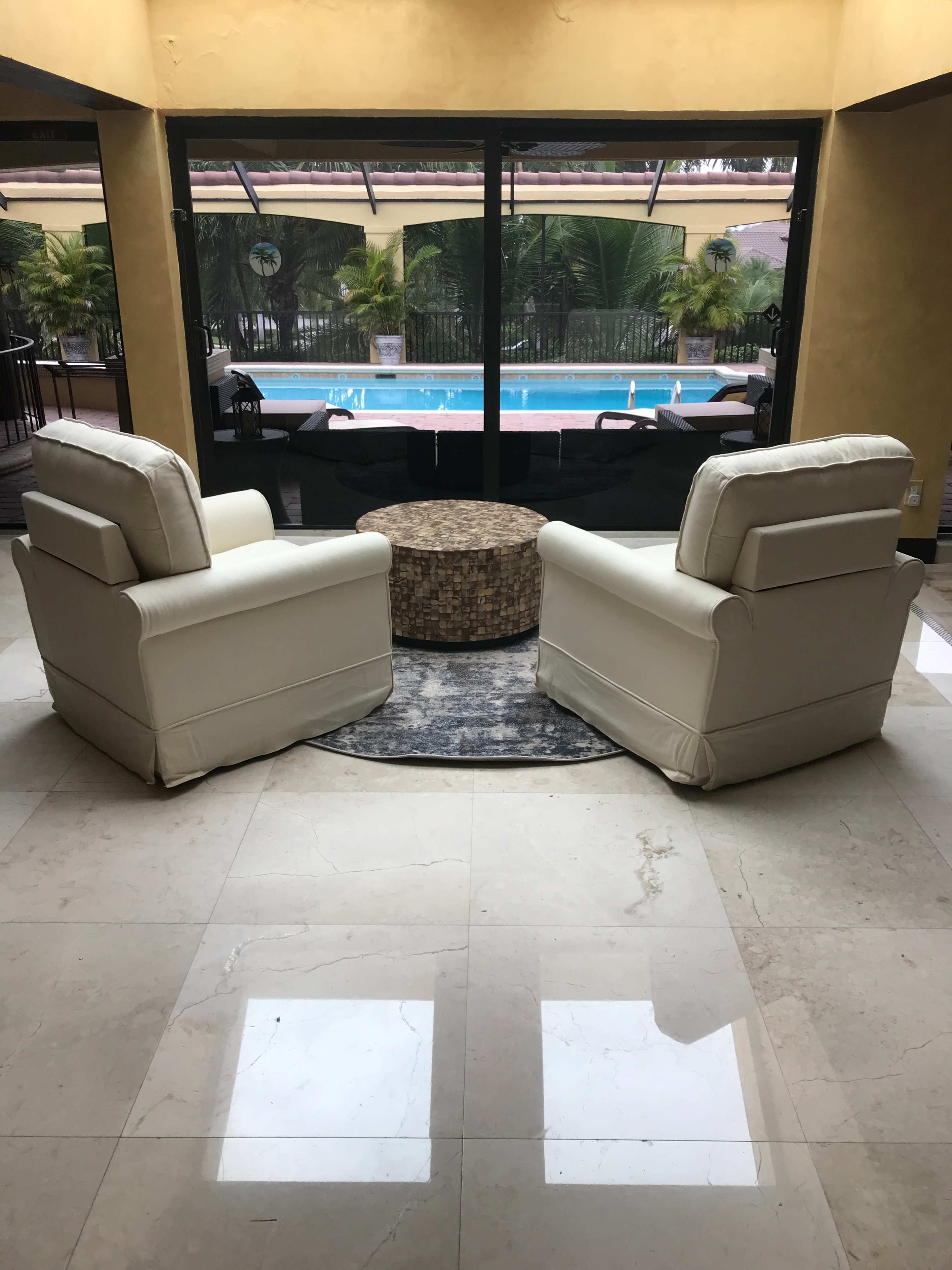 Two white armchairs face a round coffee table, set on a marble floor, with a view of a swimming pool through large glass doors.
