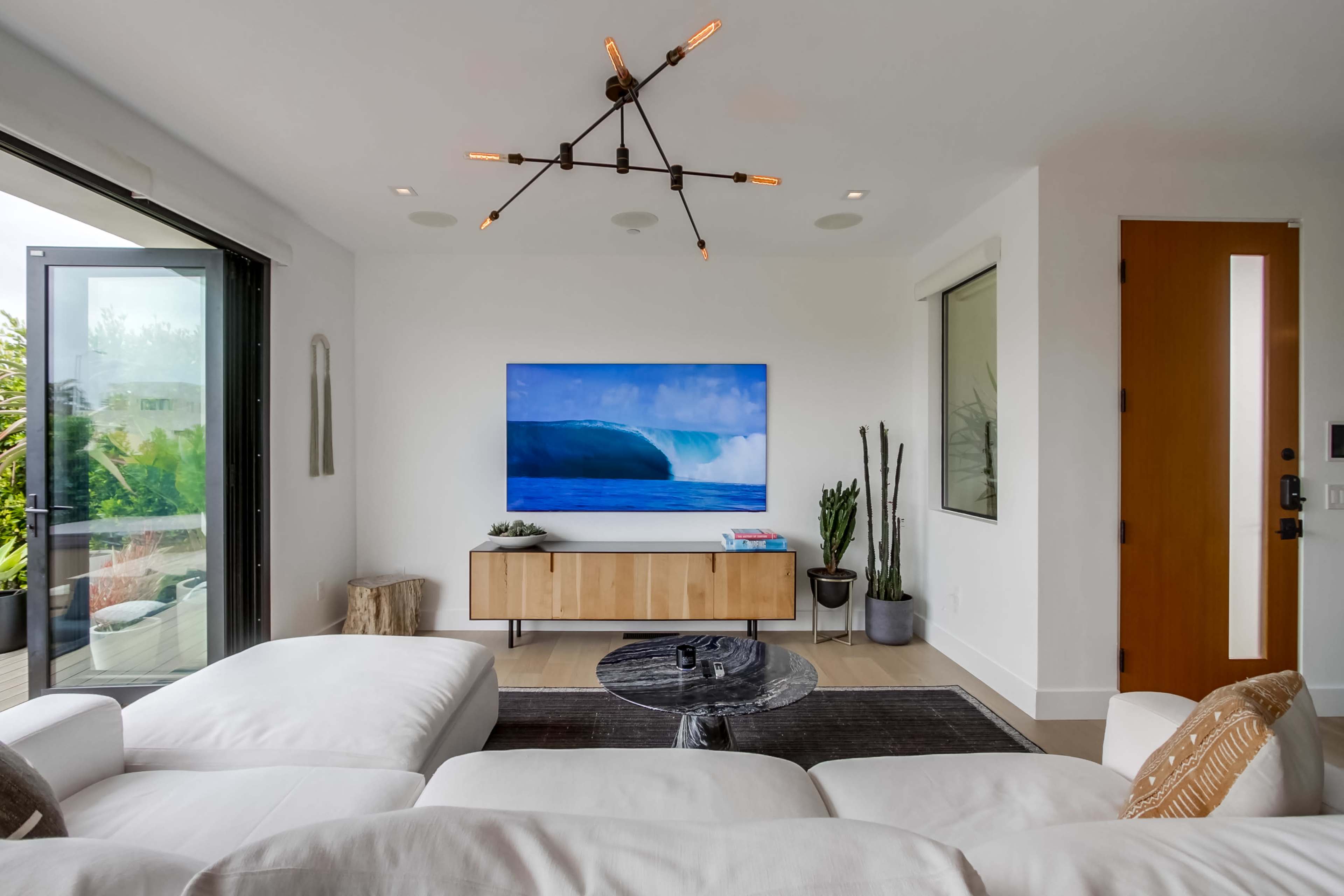 A modern living room features a white sectional sofa, a minimalist coffee table, a large wall-mounted TV displaying a scenic image, and natural light streaming through glass doors.