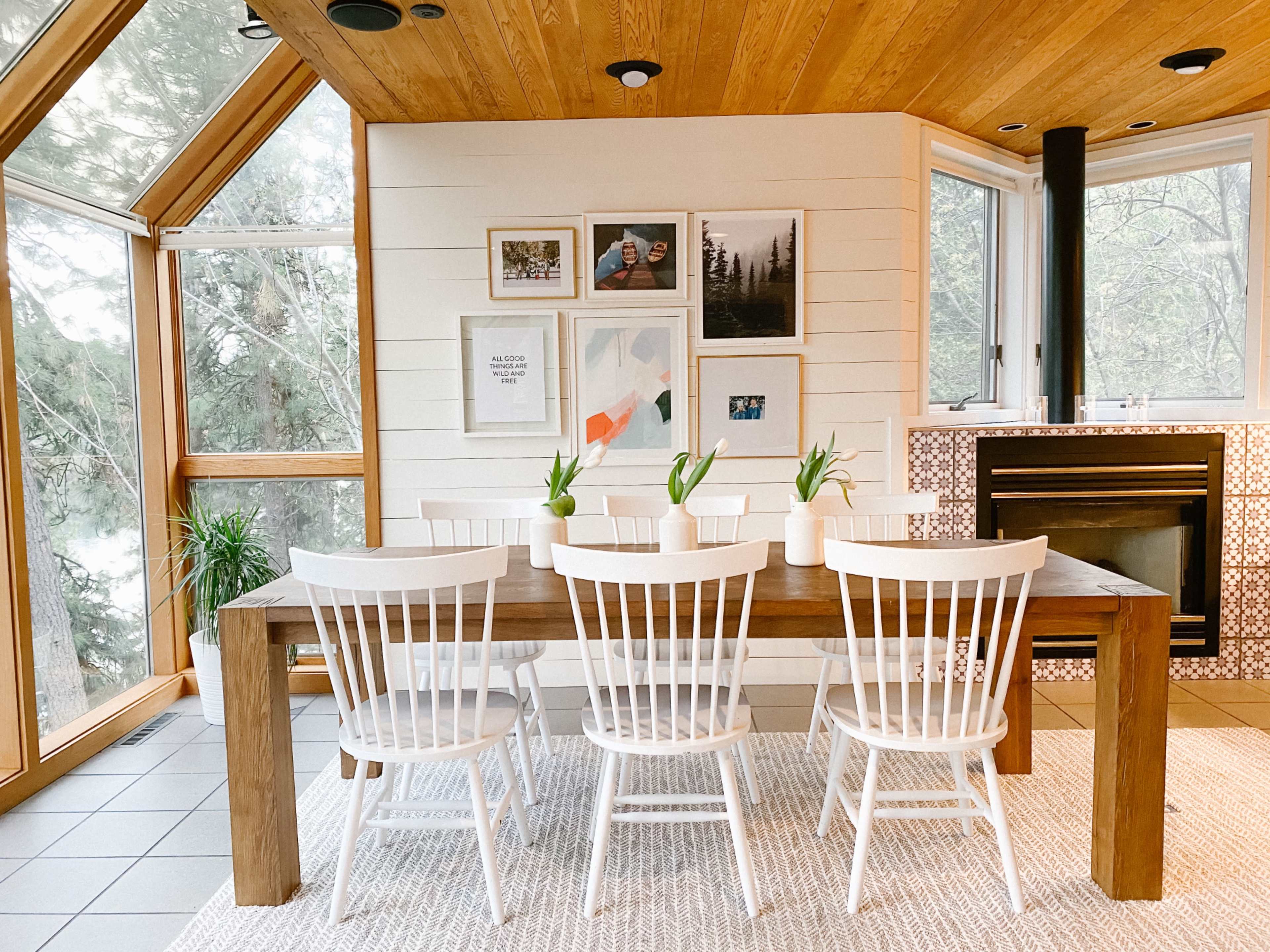 A wooden dining table surrounded by white chairs is positioned in a room with large windows and framed artwork on the walls.