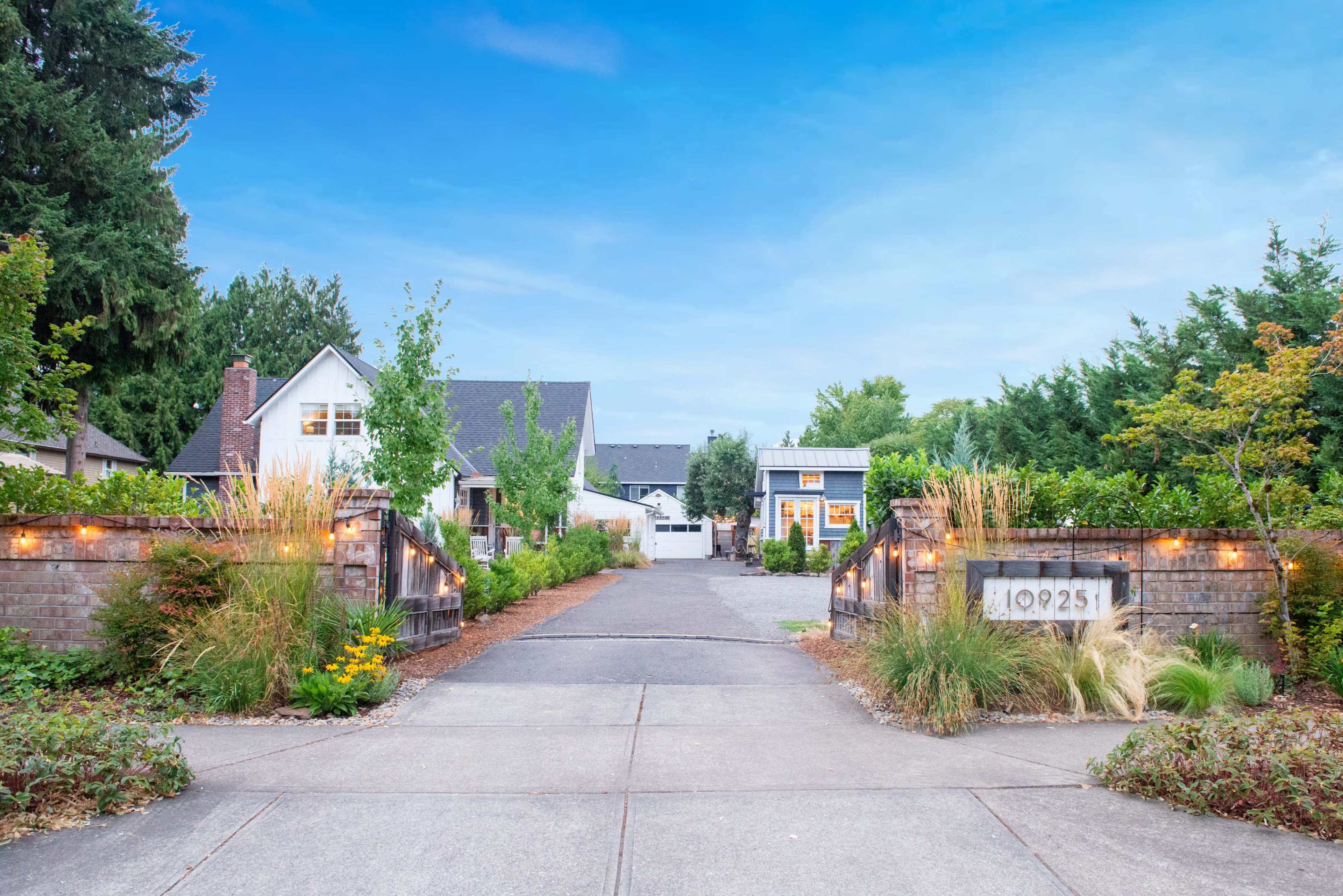 The image shows a residential driveway lined with trees and decorative lights, leading to multiple houses set against a clear blue sky.