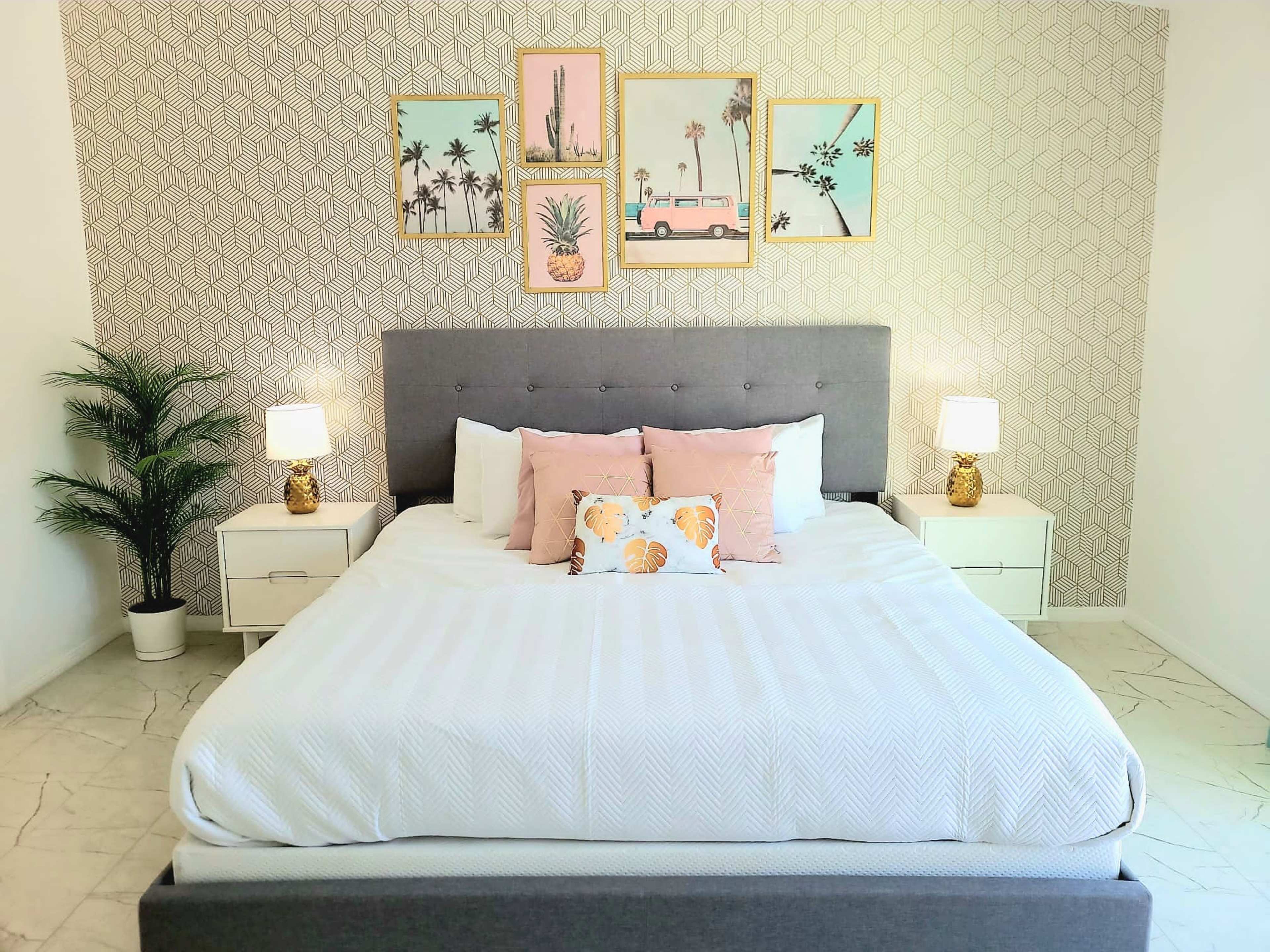 The image displays a neatly arranged bedroom with a gray upholstered bed, pink accent pillows, bedside tables with lamps, and framed artwork featuring tropical themes on the wall behind it.
