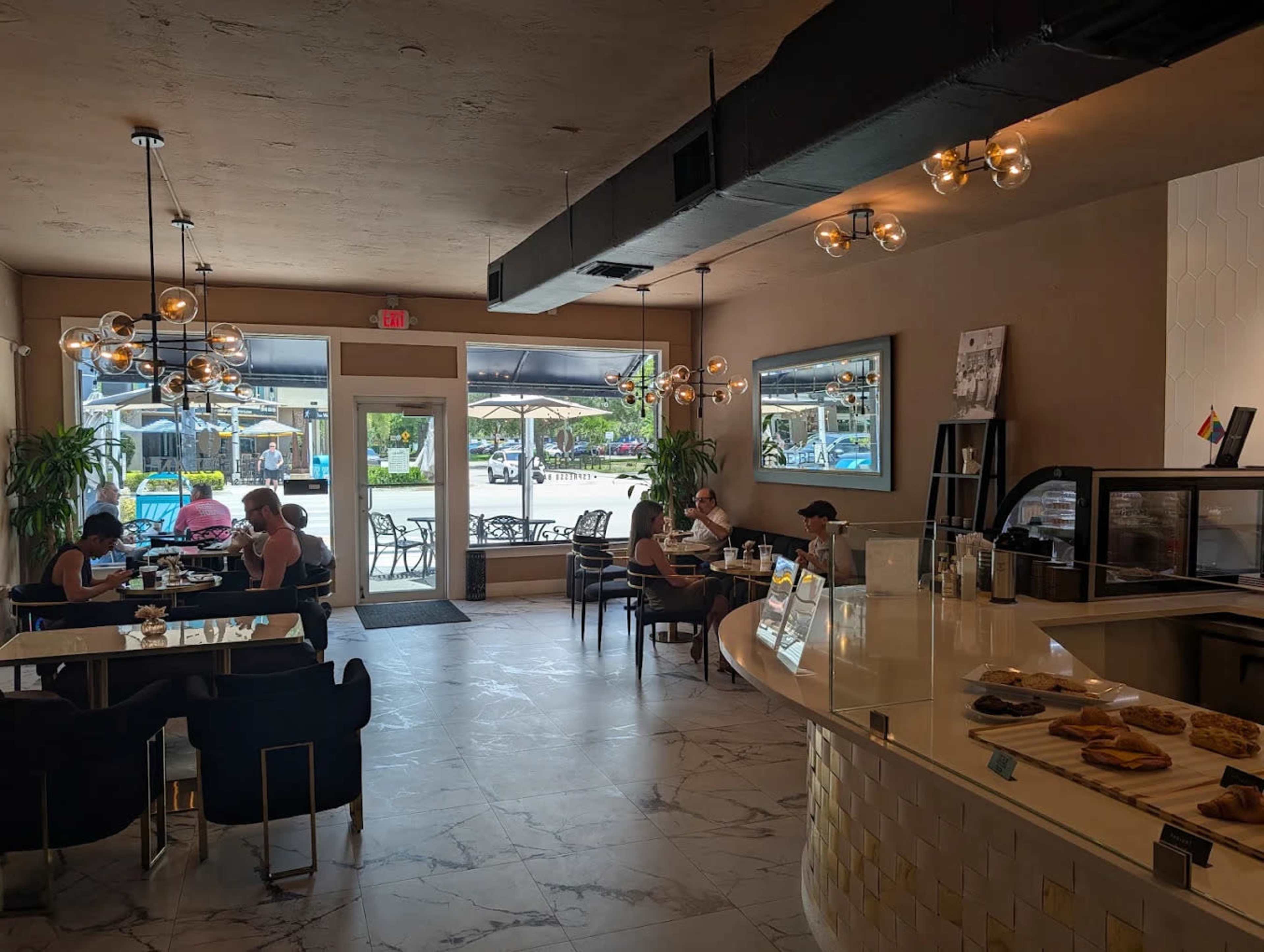 The image shows a modern café interior with patrons seated at tables, a counter displaying pastries, and large windows allowing natural light to enter.