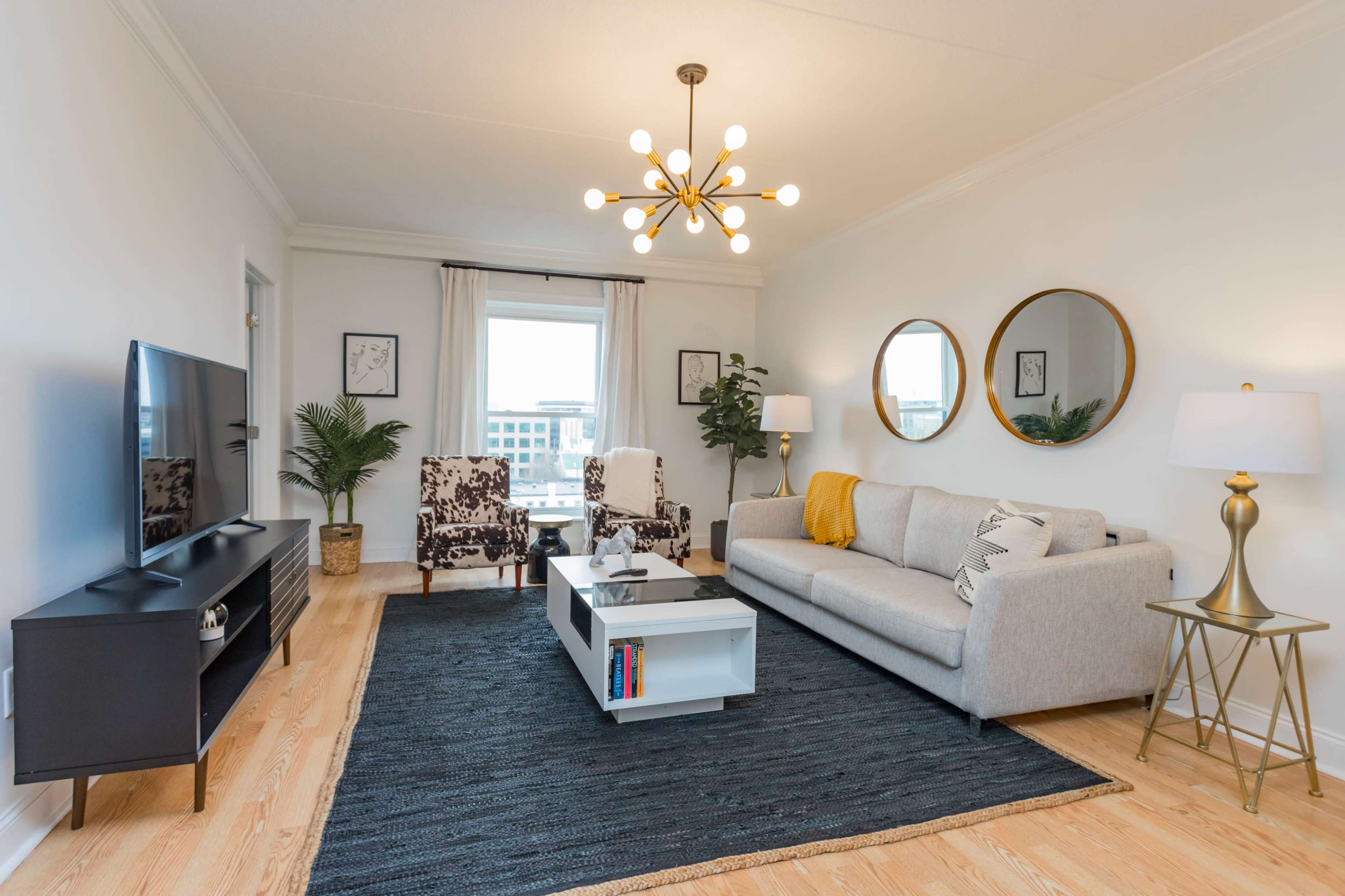 A modern living room features a gray sofa, a black TV stand, round mirrors, and patterned chairs, with a light fixture overhead and a large window illuminating the space.