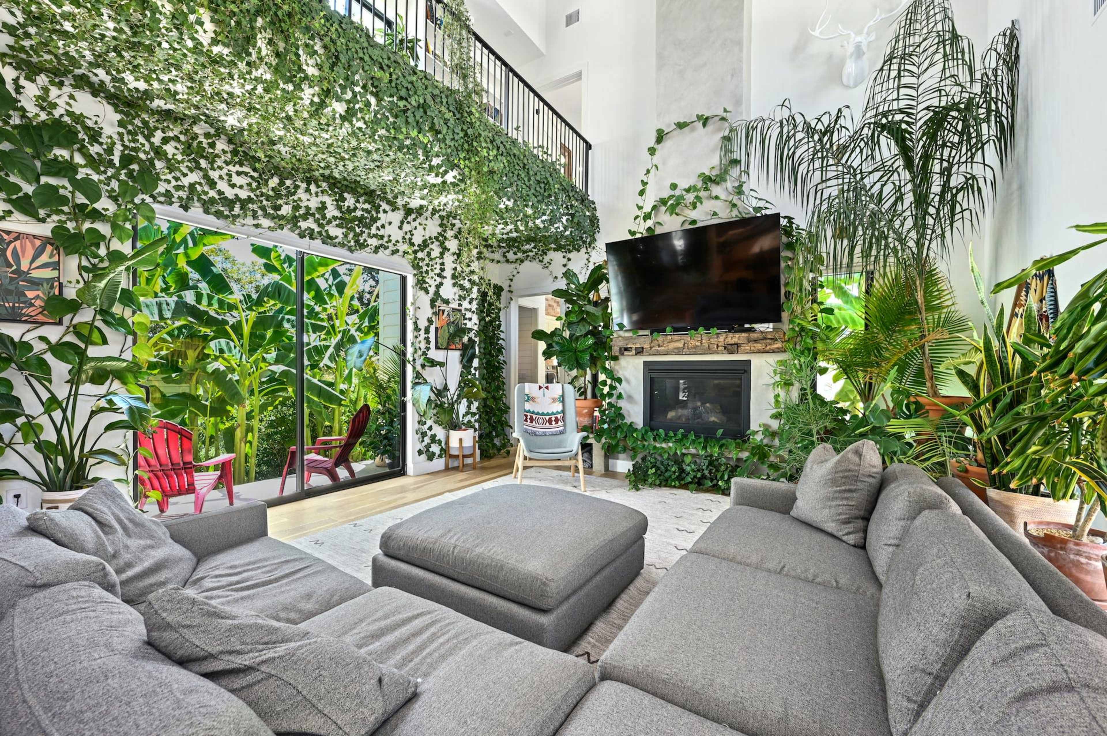 A spacious living room filled with lush green plants, featuring a large sectional sofa, a mounted television, and expansive windows opening to a patio.