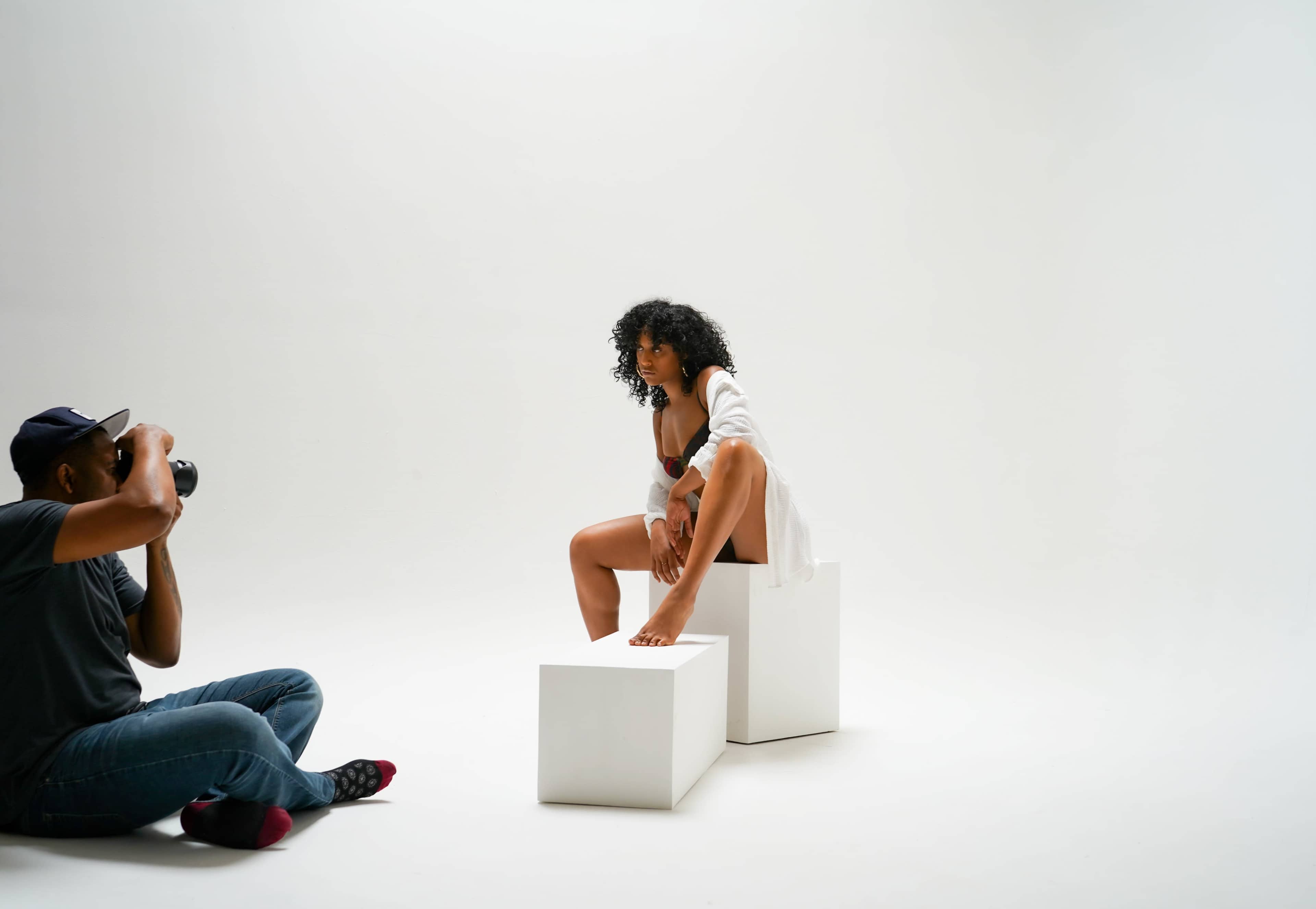 A photographer captures a model posing on a white block in a minimalist studio setting.