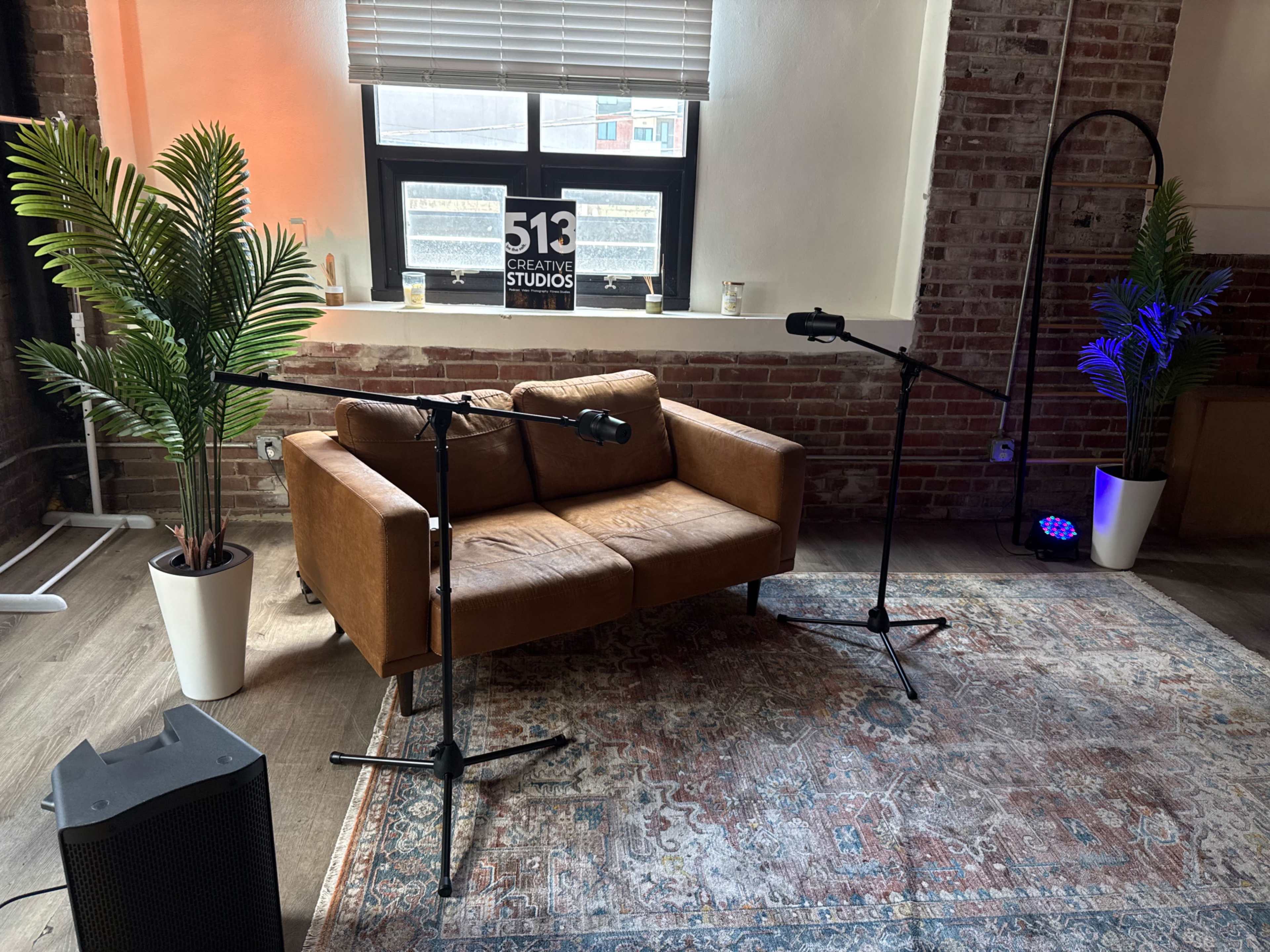 The image shows a cozy seating area with a brown leather sofa, two microphones on stands, and potted plants in a room with exposed brick walls and a large window.