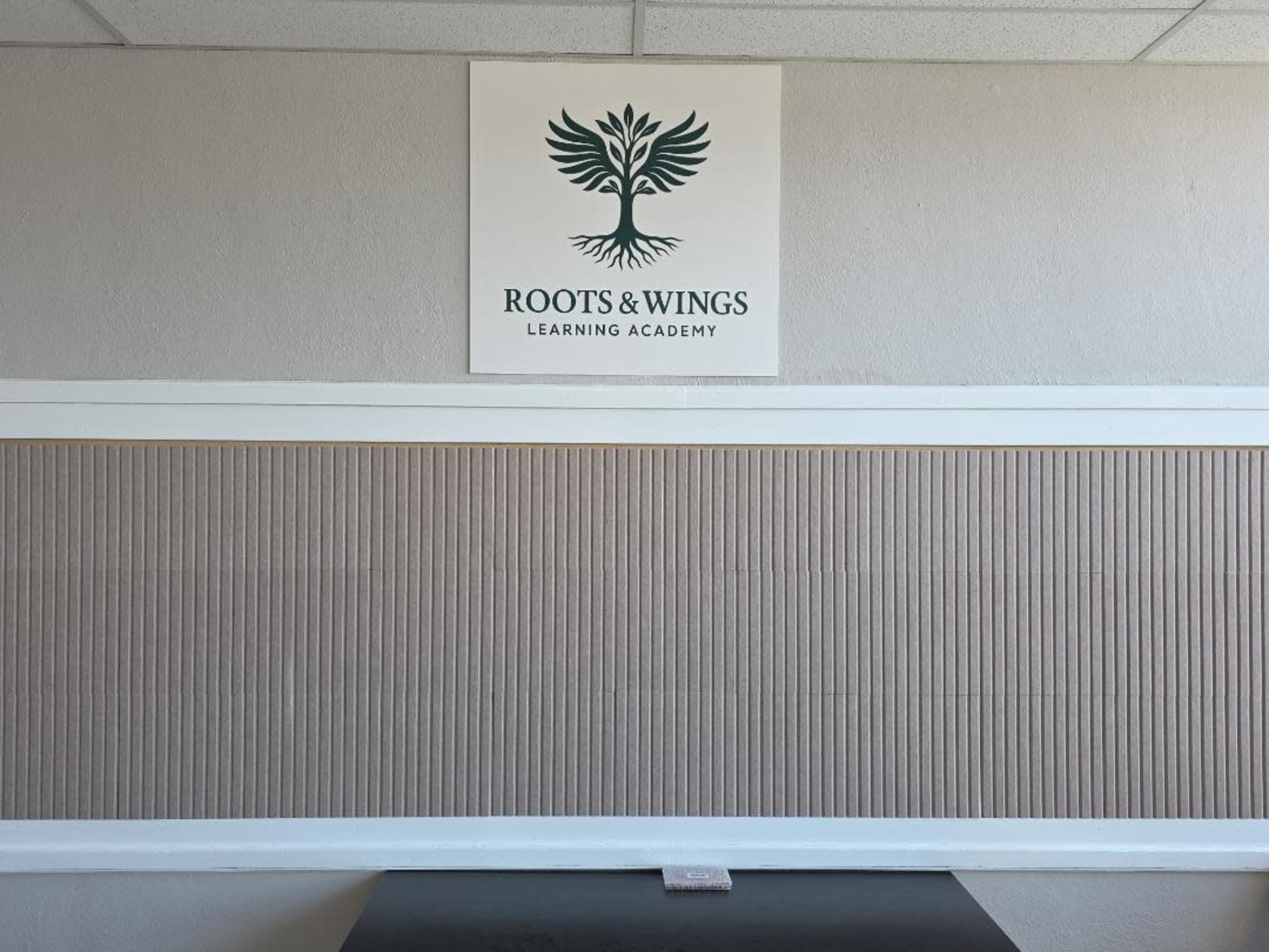 The image shows the interior wall of a building featuring a sign for "Roots & Wings Learning Academy" above a textured panel.