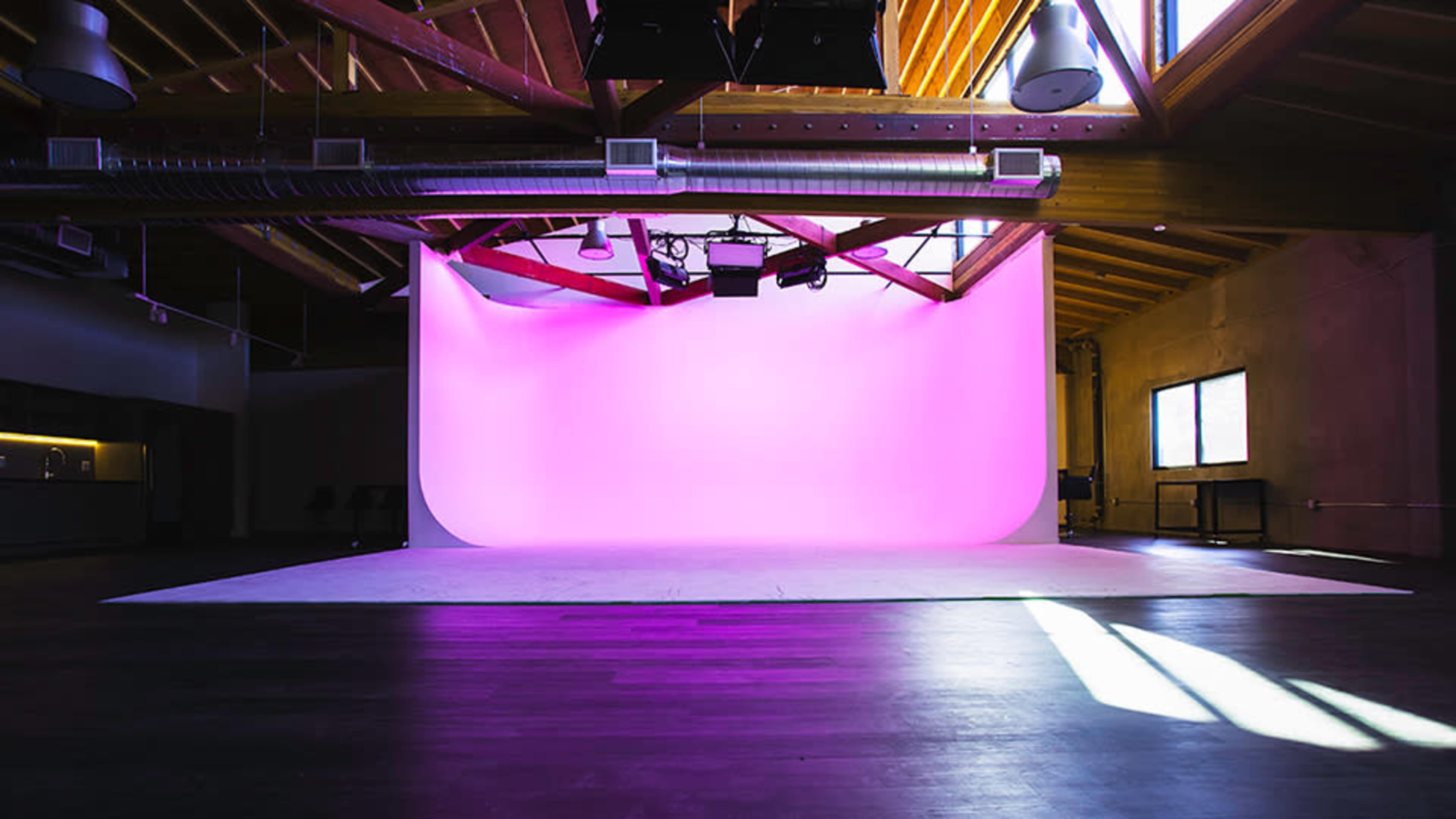 A spacious photo studio with a pink backdrop and wooden beams on the ceiling, illuminated by natural light.