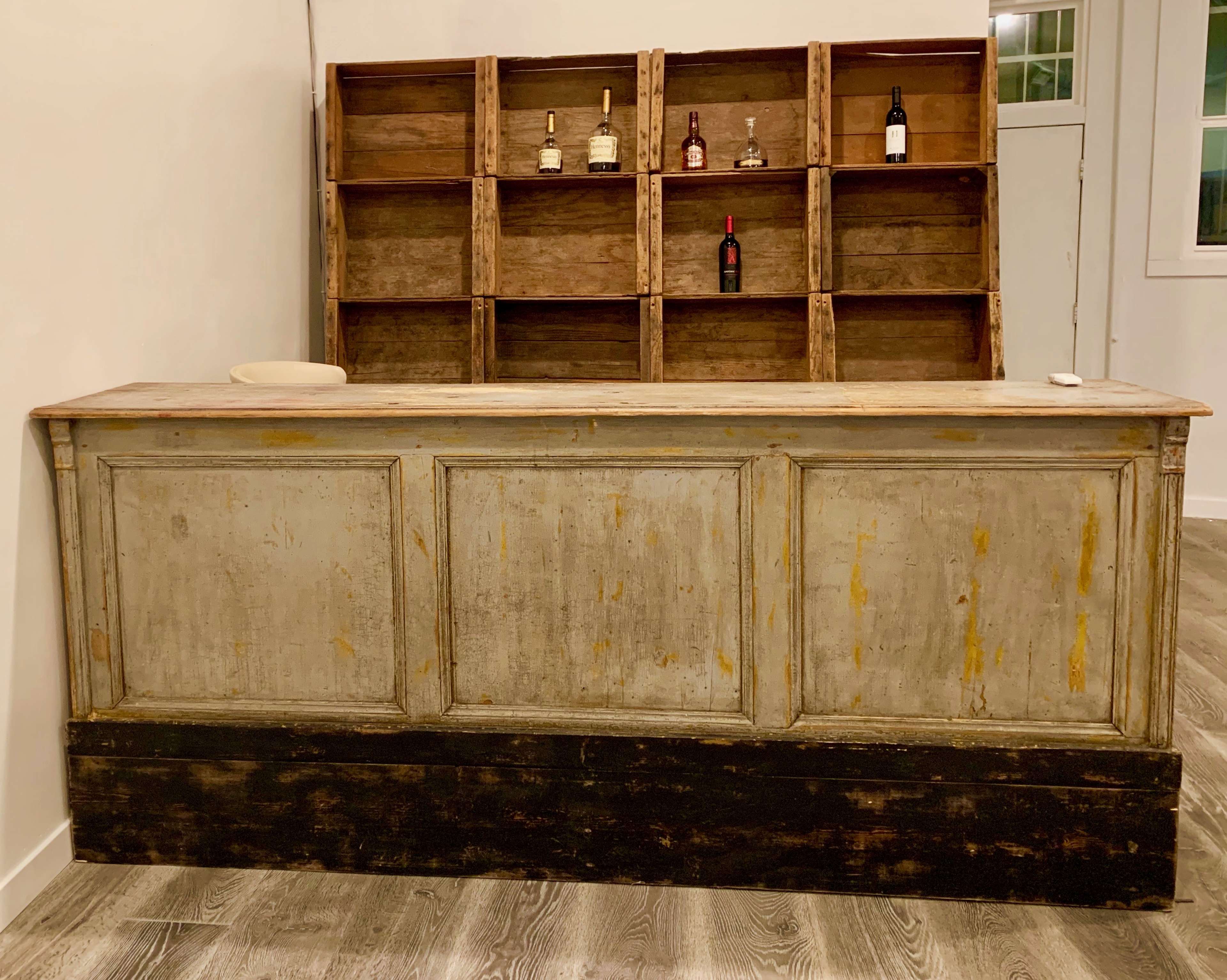 A wooden bar counter with a rustic finish, set in front of a shelf displaying several bottles of wine and liquor against a plain wall.