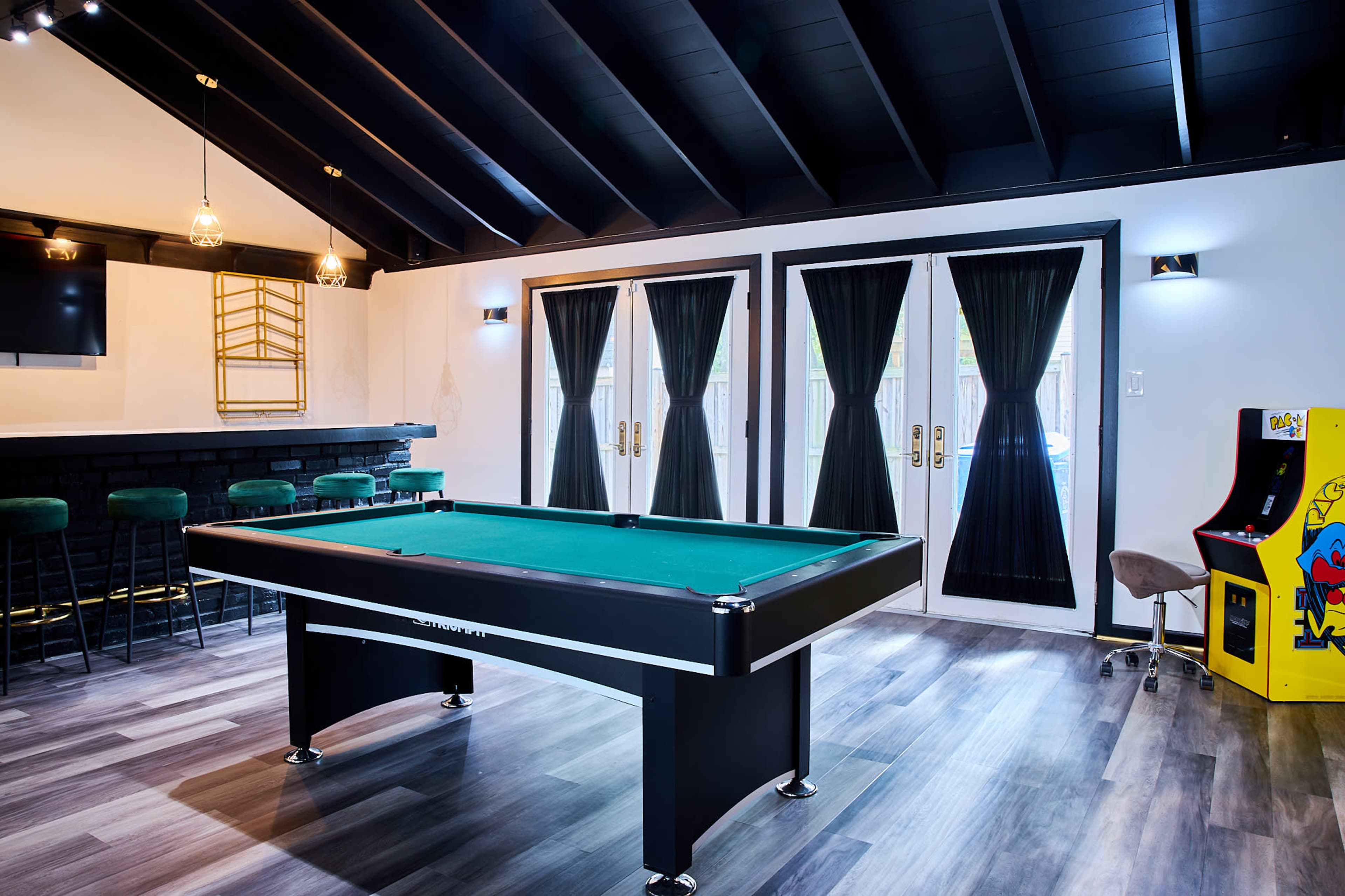 A game room with a green pool table, a bar with stools, and a classic arcade cabinet, all under a ceiling with exposed wooden beams.