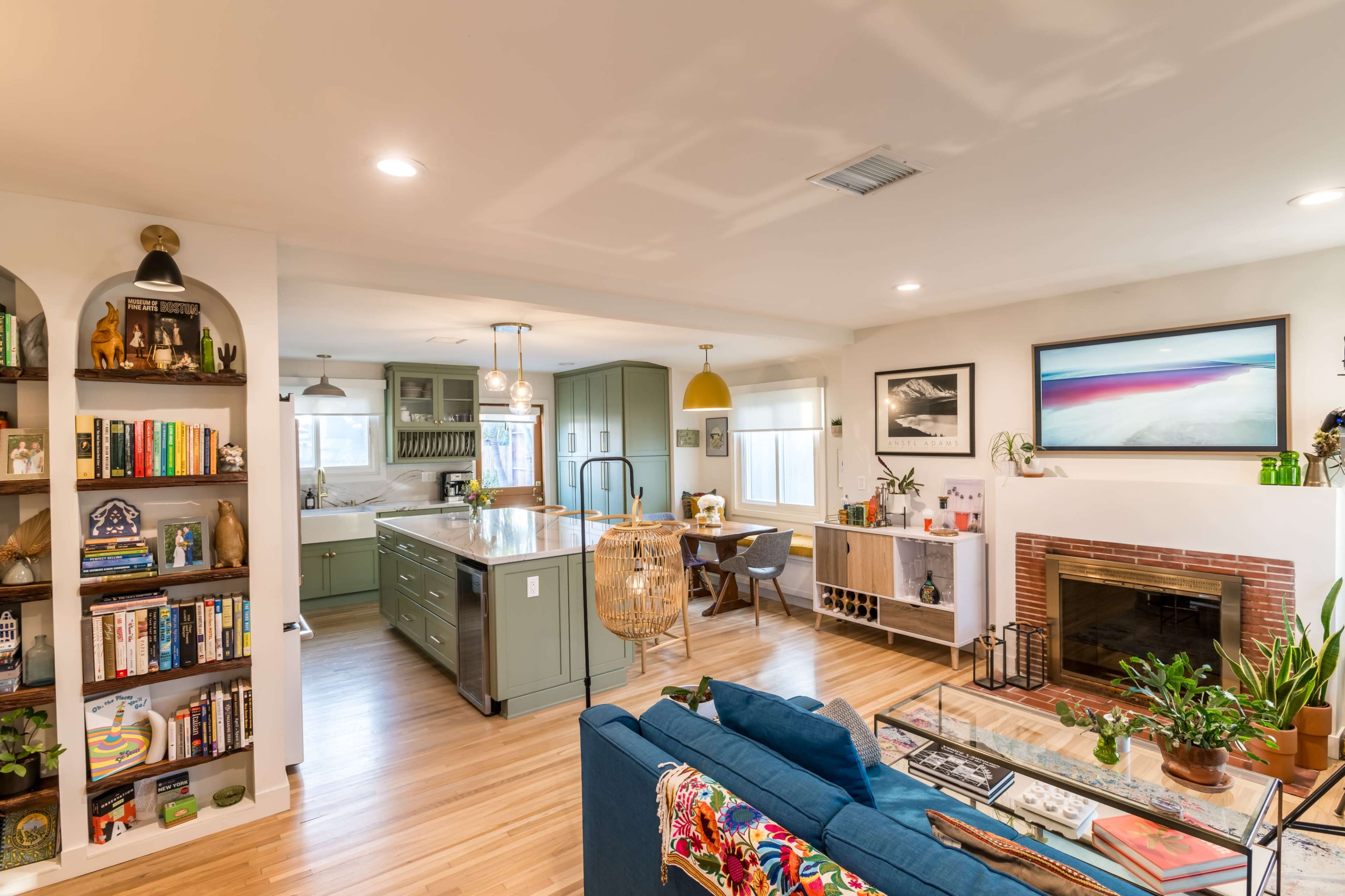 A modern living space featuring a blue sofa, a bookshelf filled with books, a kitchen with green cabinetry, and a fireplace adorned with plants.