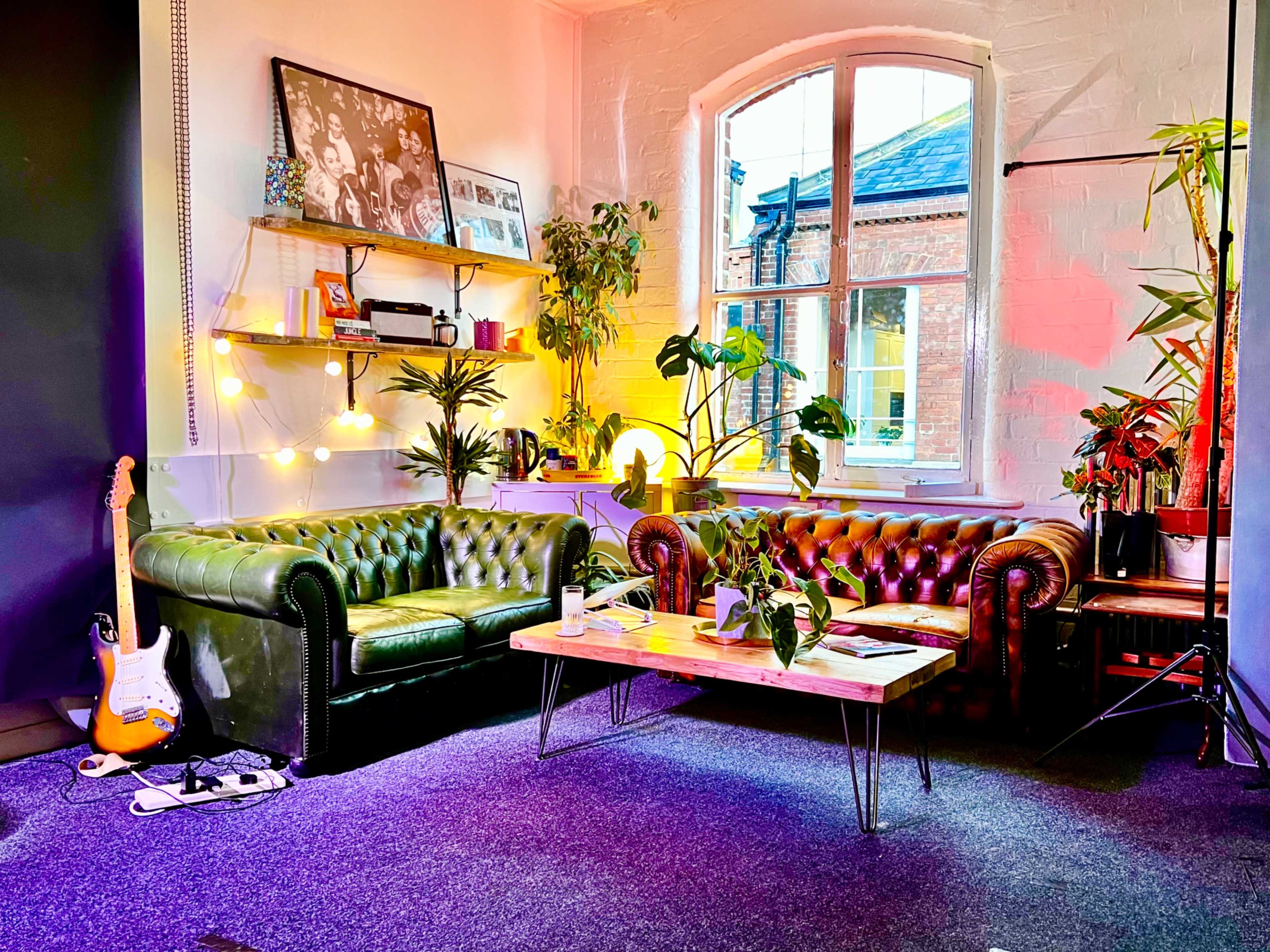 The scene features a cozy seating area with two leather sofas, a wooden coffee table surrounded by plants, and a large window providing natural light.