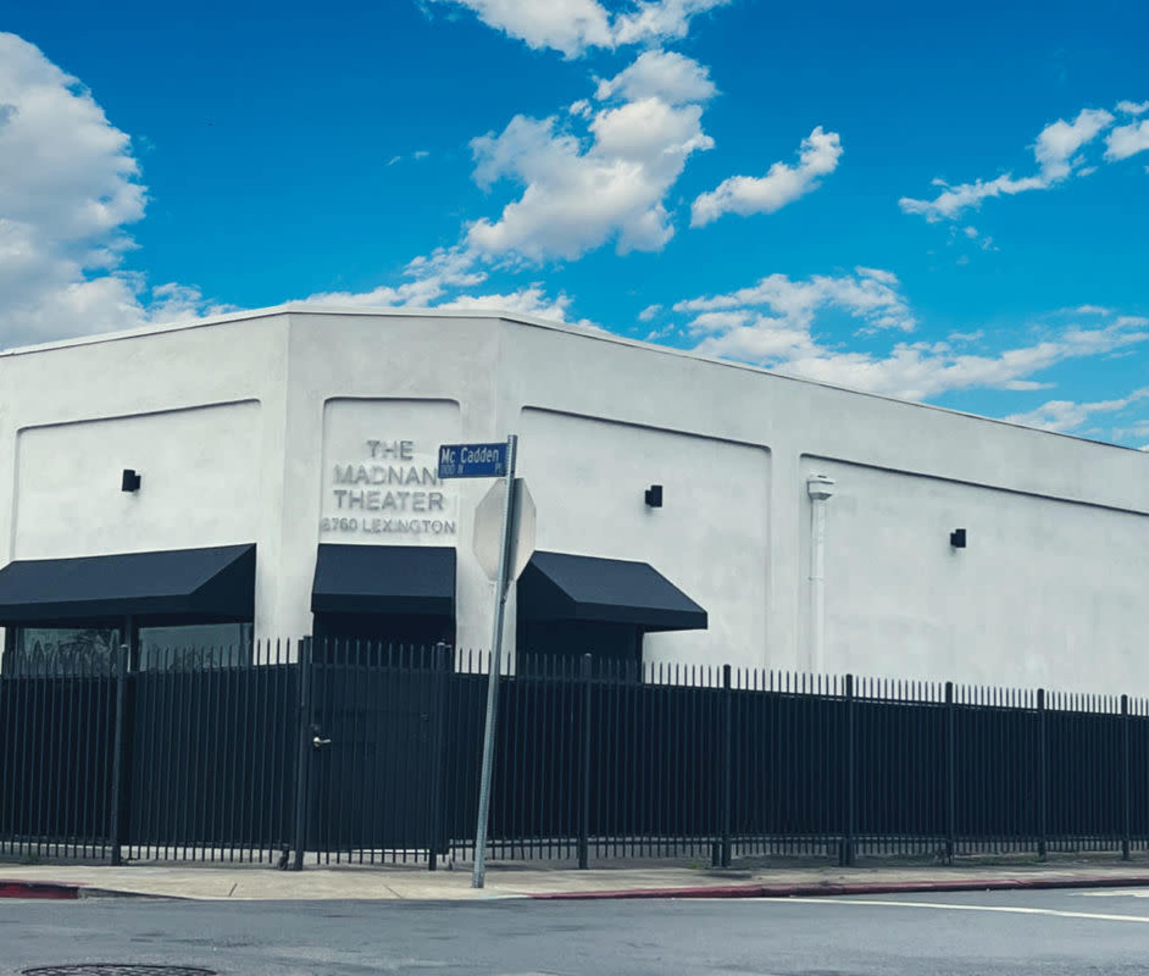 The image shows a white building with a flat roof, labeled "THE MADAM THEATER," surrounded by a black fence against a partly cloudy blue sky.