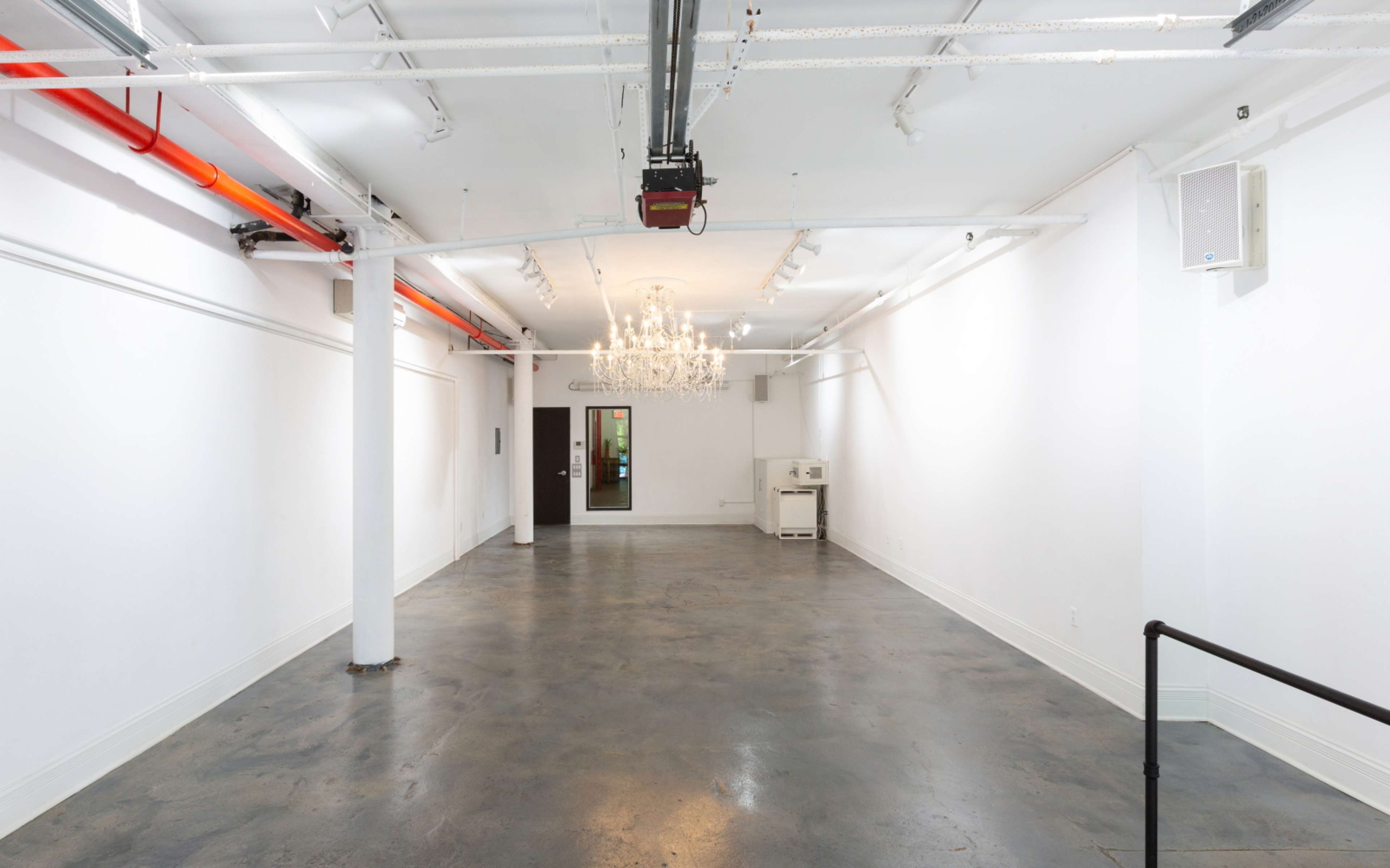 A spacious, empty room with concrete flooring, white walls, and a chandelier hanging from the ceiling.