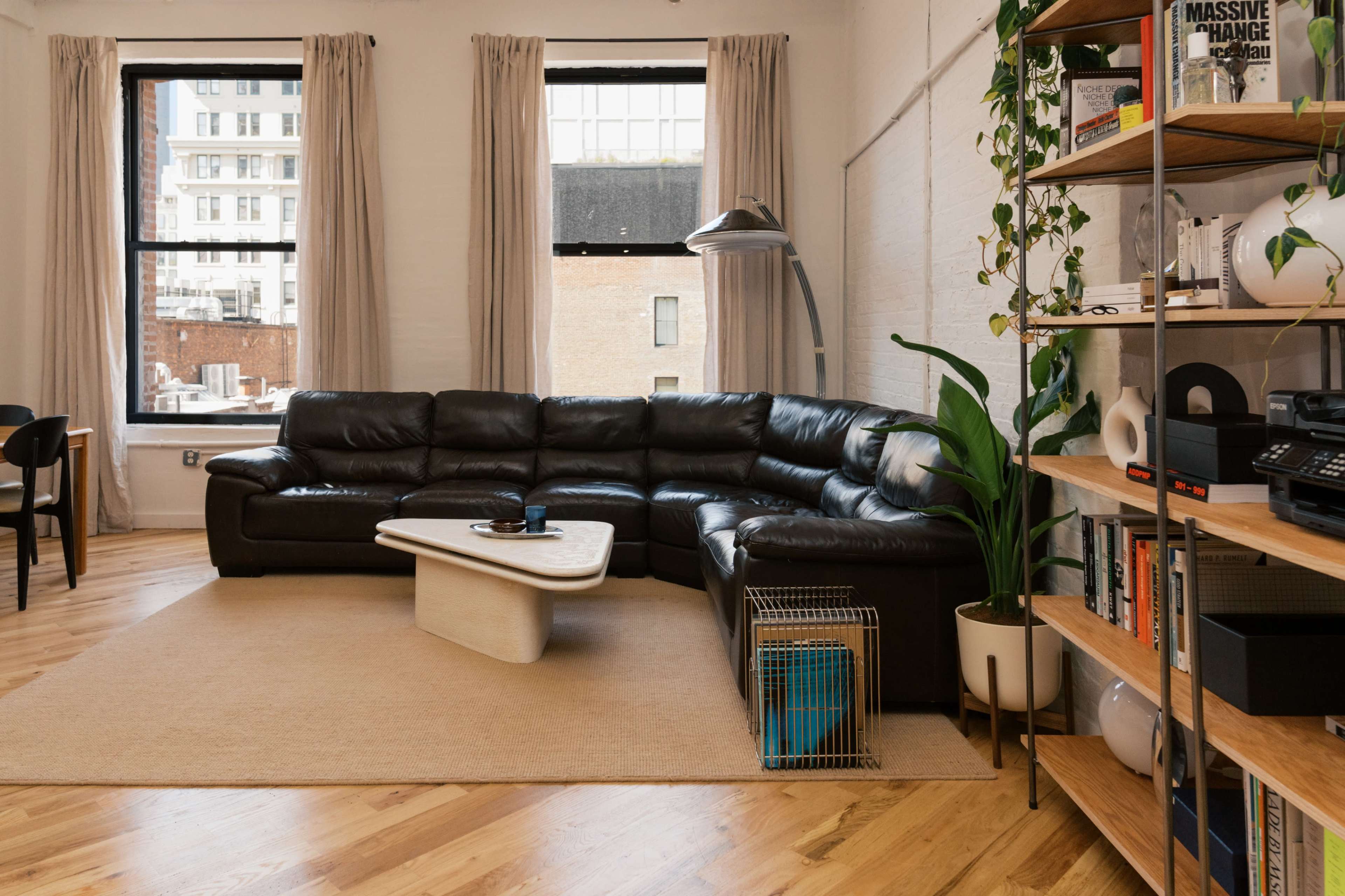 The image shows a spacious living room featuring a large black sectional sofa, a light-colored coffee table, and potted plants, with windows letting in natural light.