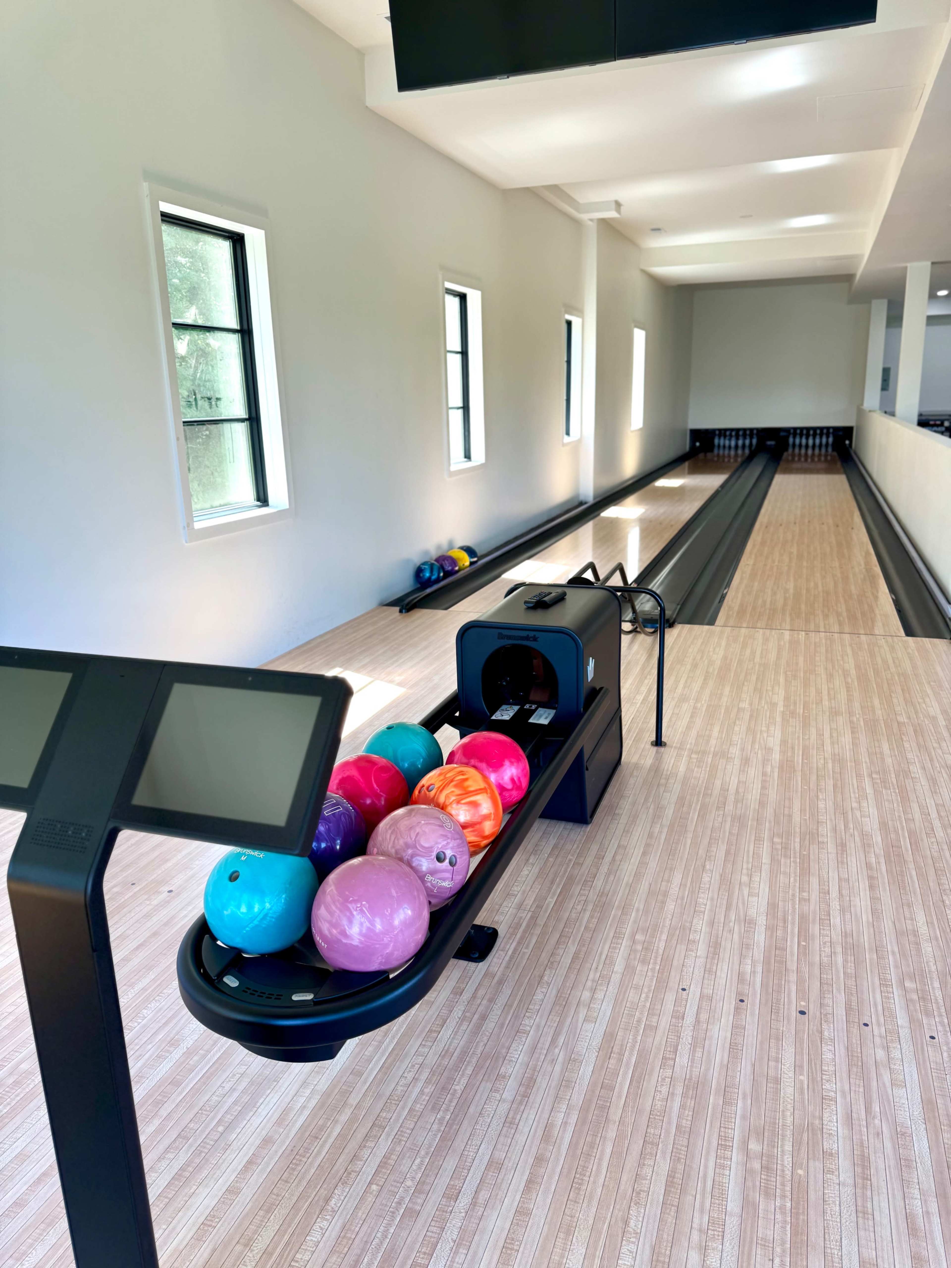 The image shows a bowling alley with a polished wooden lane, a ball return system, and several colorful bowling balls on a rack.