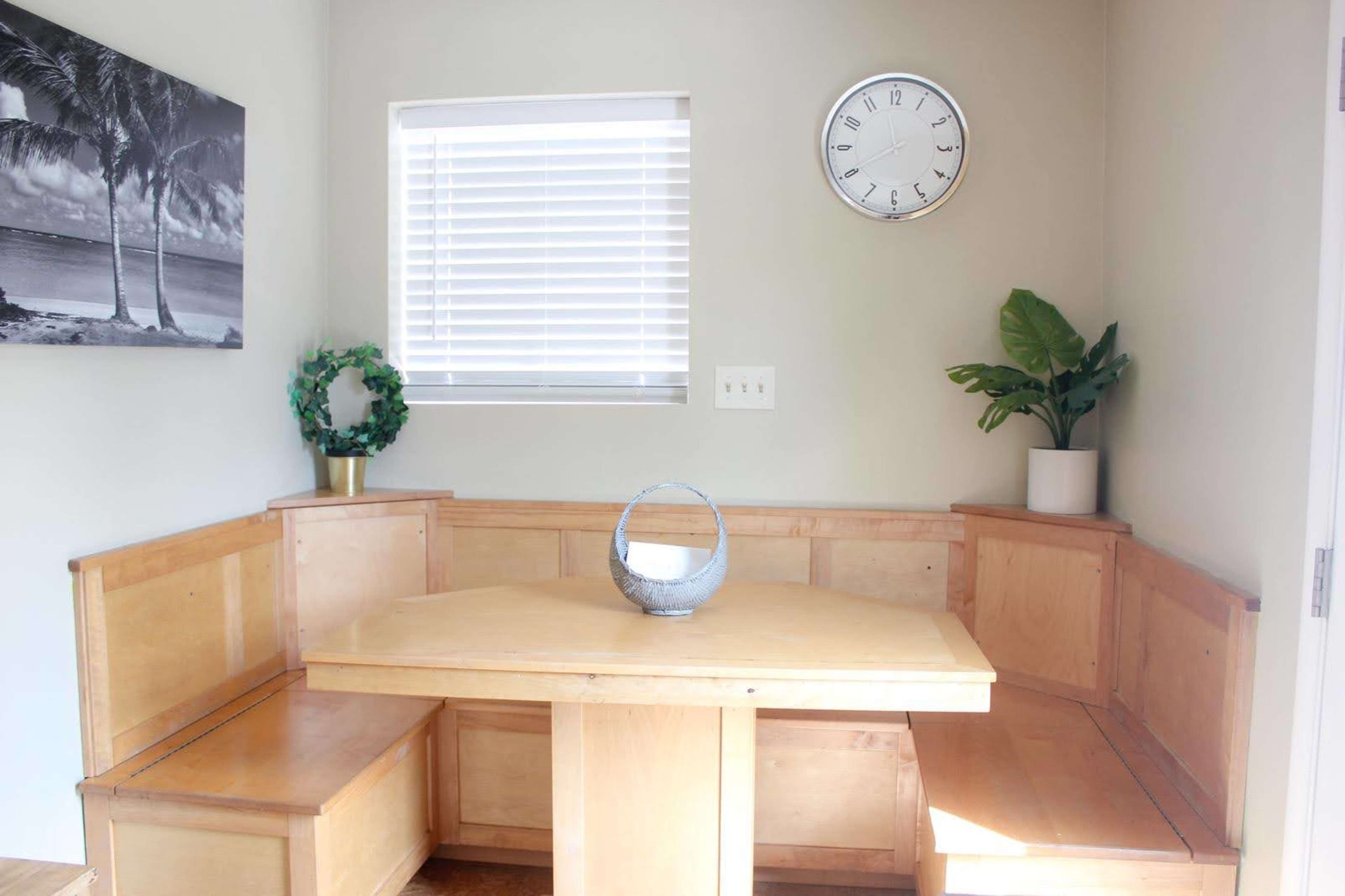 A corner dining nook with wooden seating, a round table, a decorative basket, and two plants next to a window.