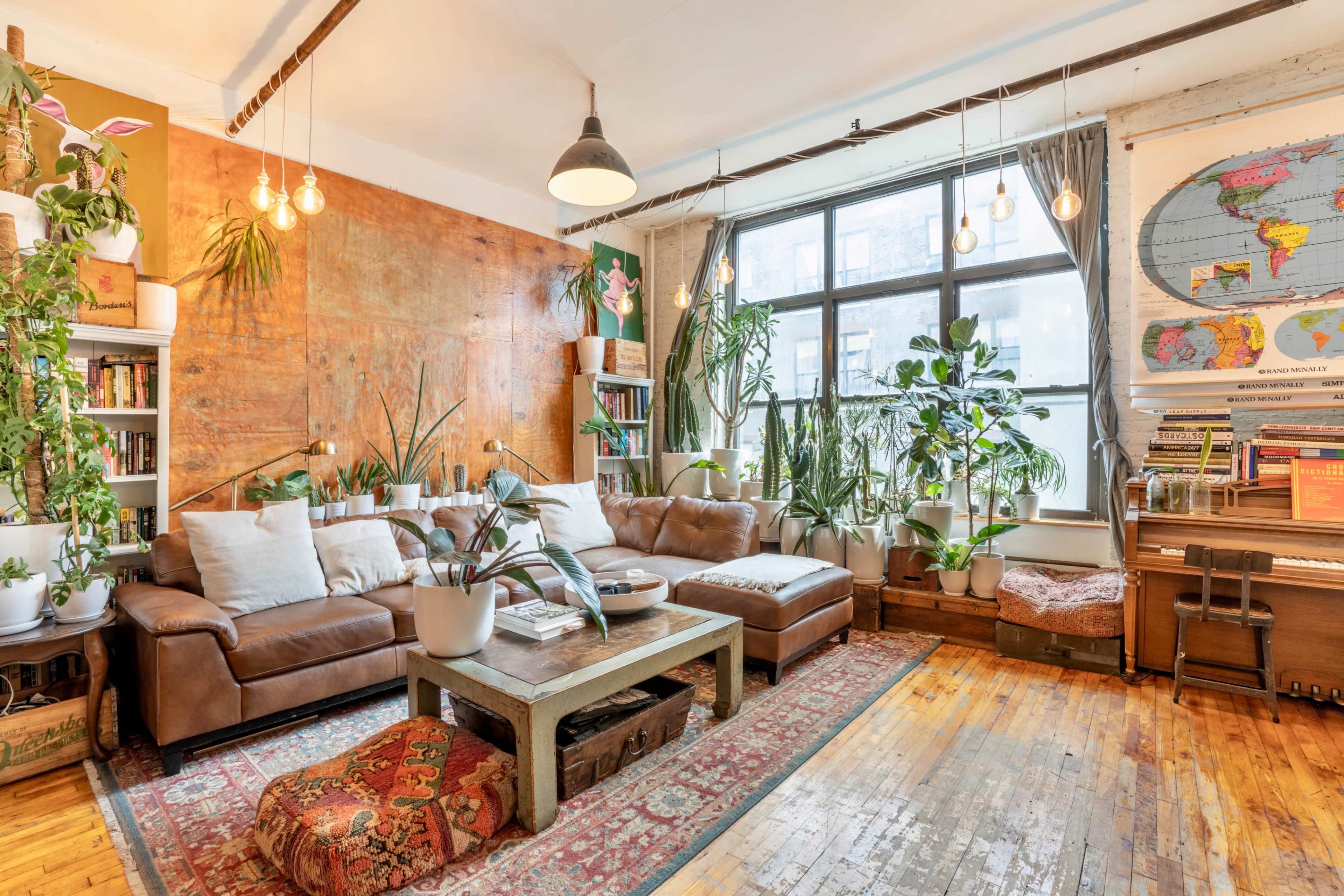 A cozy living room with a large leather sectional sofa, a coffee table, houseplants in various pots, and a piano against the wall adorned with a world map.