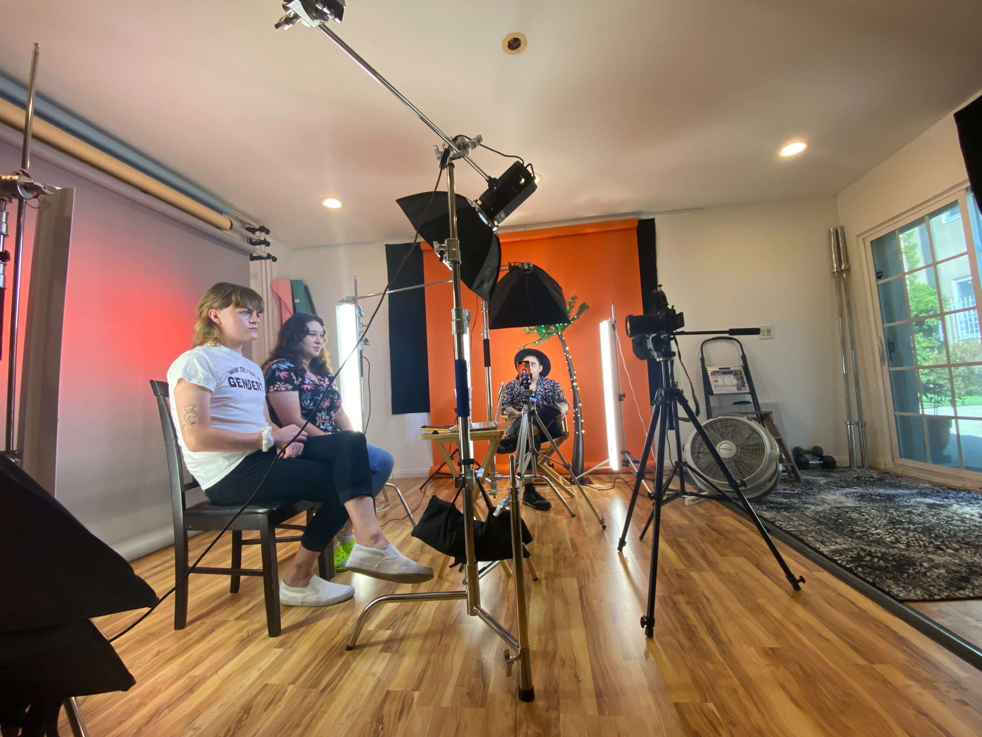 Two women are seated on a set with multiple lights and a camera, while a third person sits in front of them, all set against an orange backdrop.
