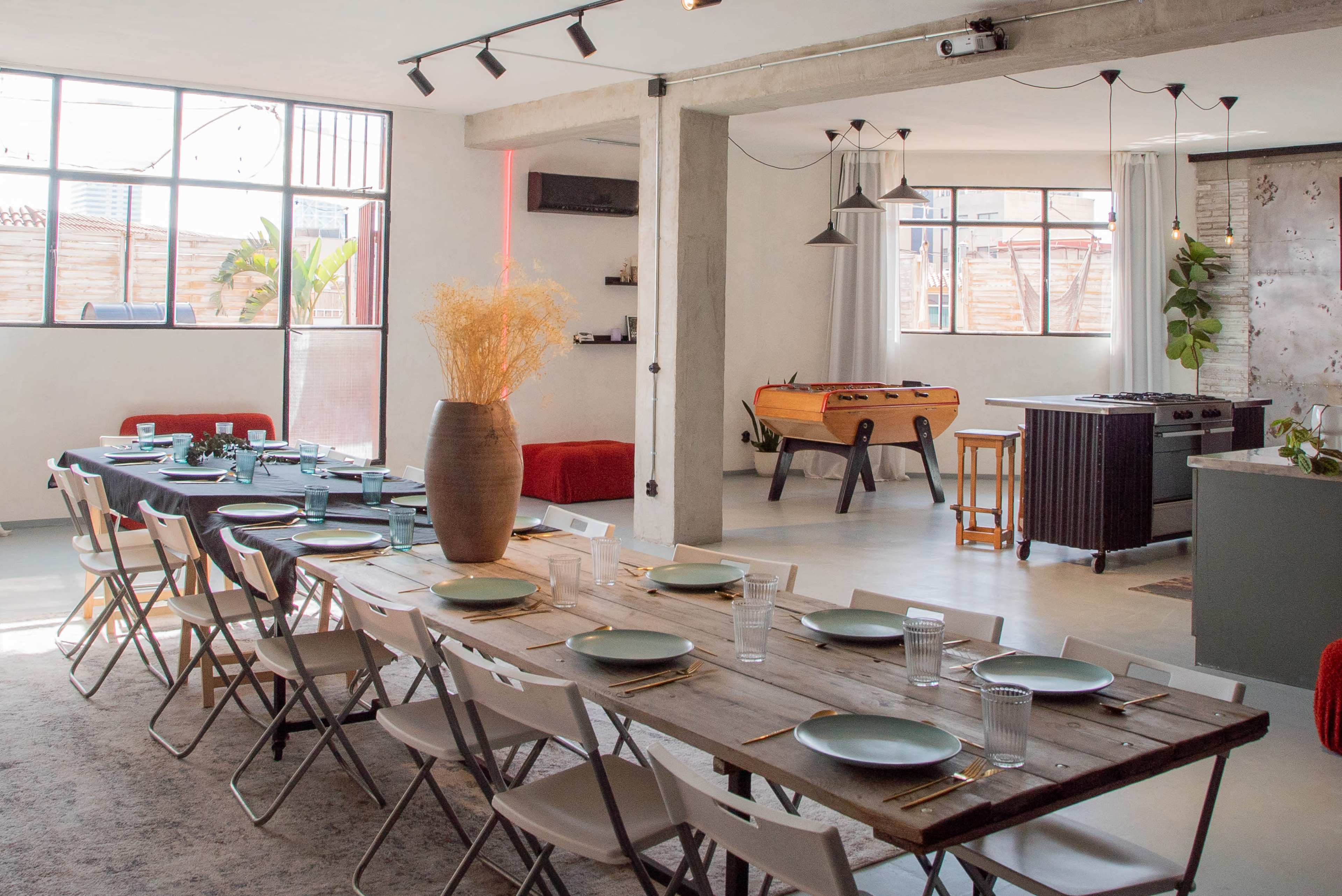 The image shows a modern, open-plan dining area with a long wooden table set for a meal, surrounded by folding chairs, and featuring a foosball table in the background.