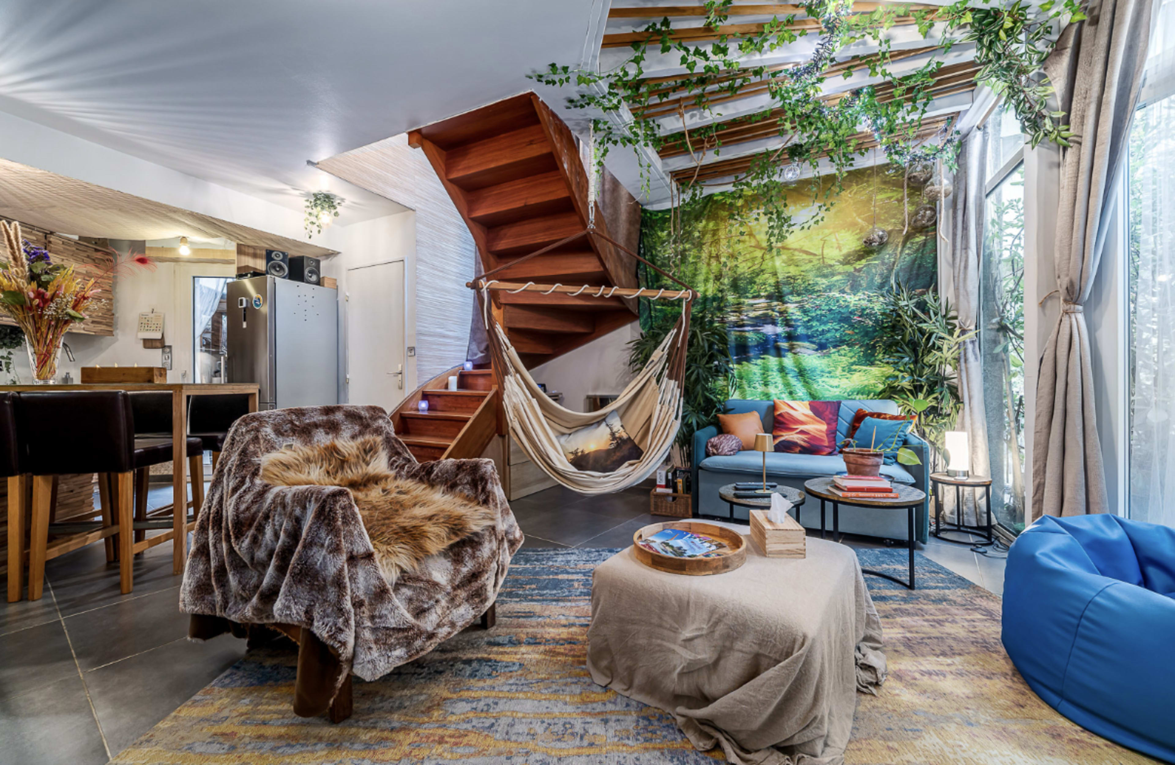 The image shows a cozy living space featuring a hammock, a staircase, and a colorful arrangement of furniture and plants.
