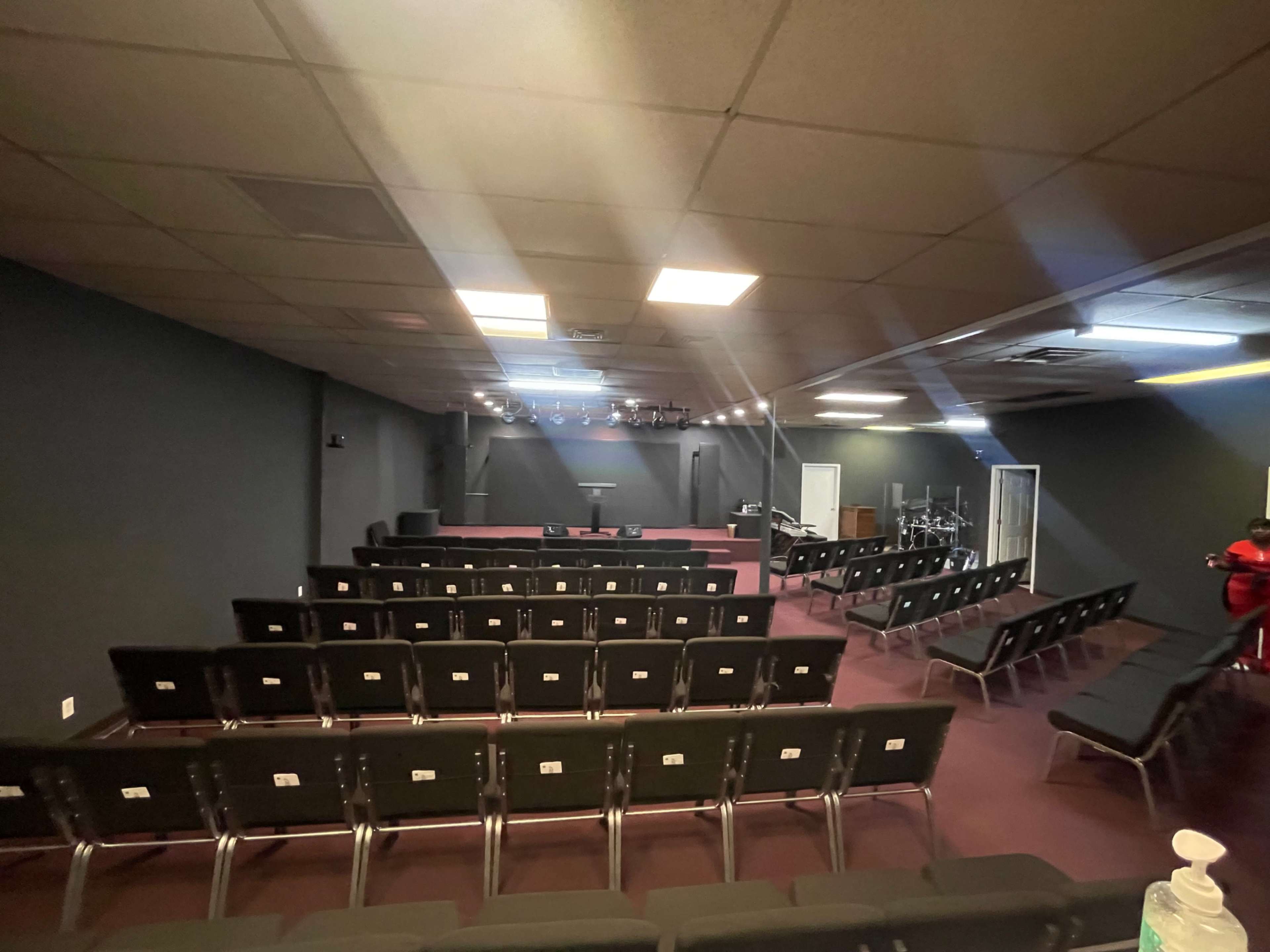 The image shows a large, empty room arranged with rows of black chairs facing a stage area with spotlights above.