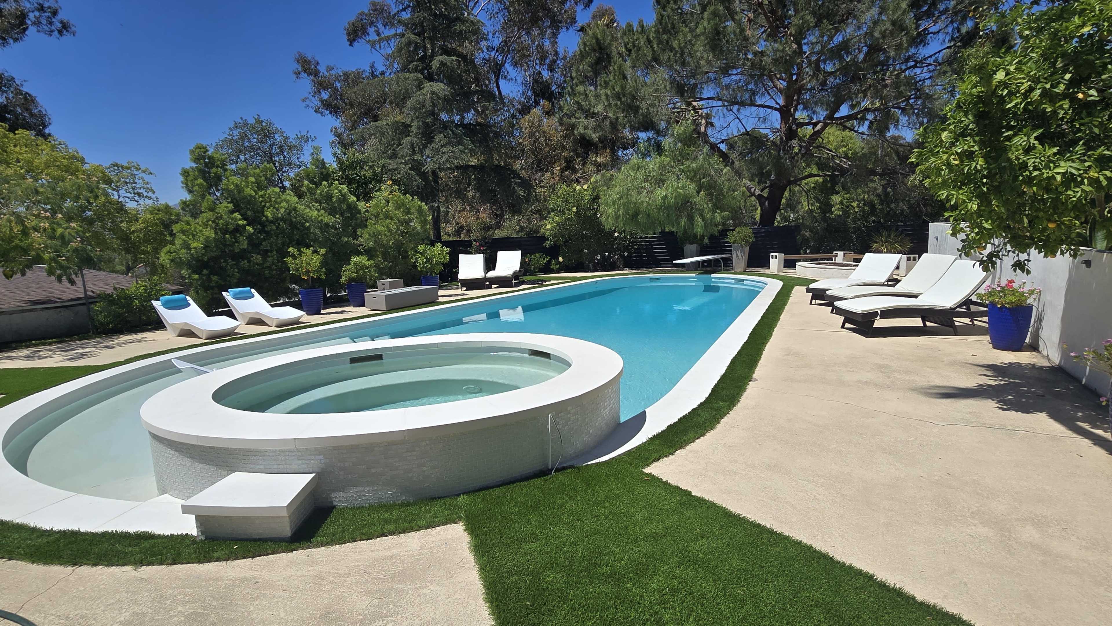 The image shows a rectangular swimming pool with a circular hot tub adjacent to it, surrounded by lounge chairs and greenery in a sunny outdoor setting.