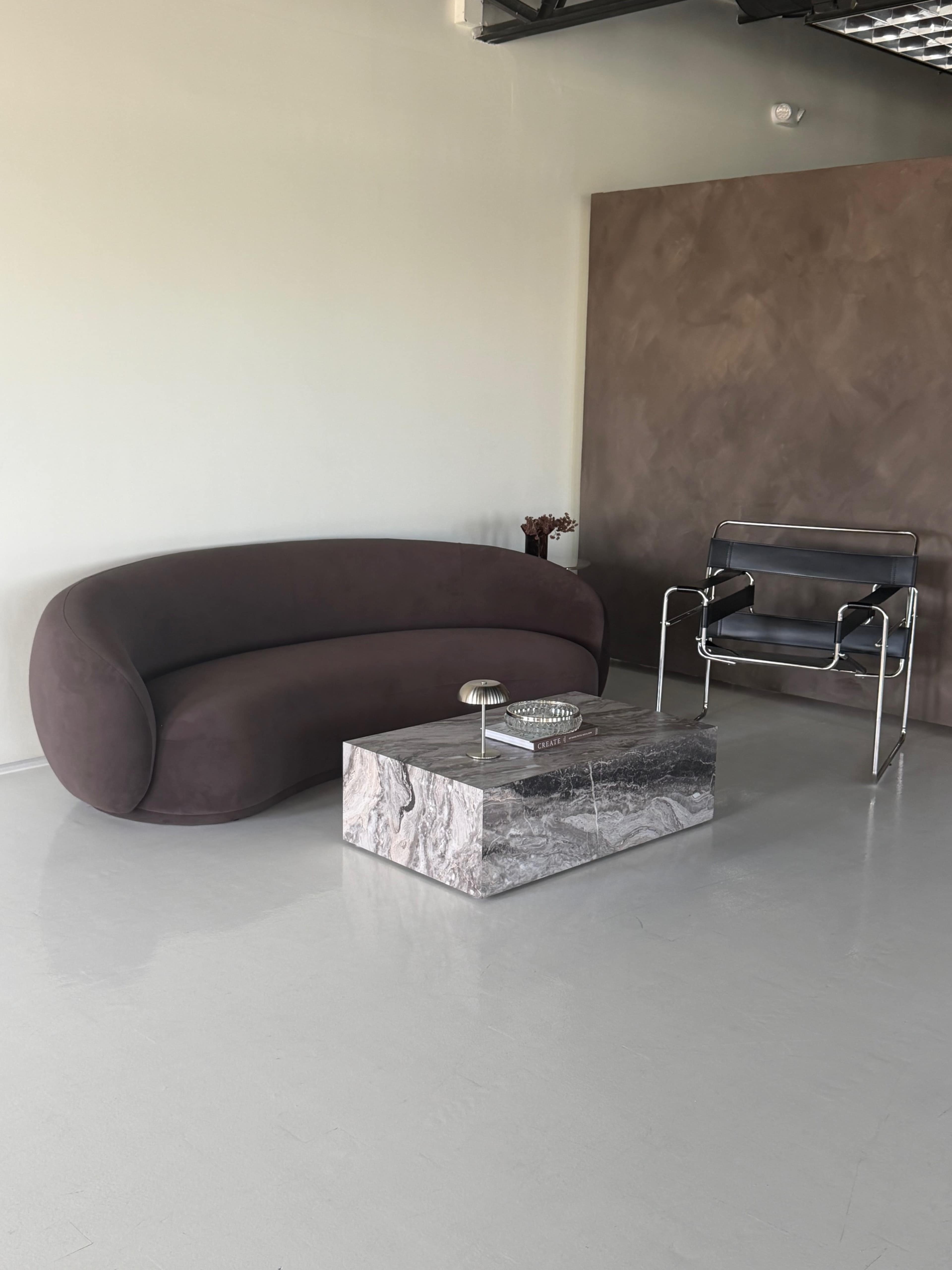 The image features a modern, curved brown sofa paired with a marble coffee table and a sleek metal armchair in a minimalist interior space.