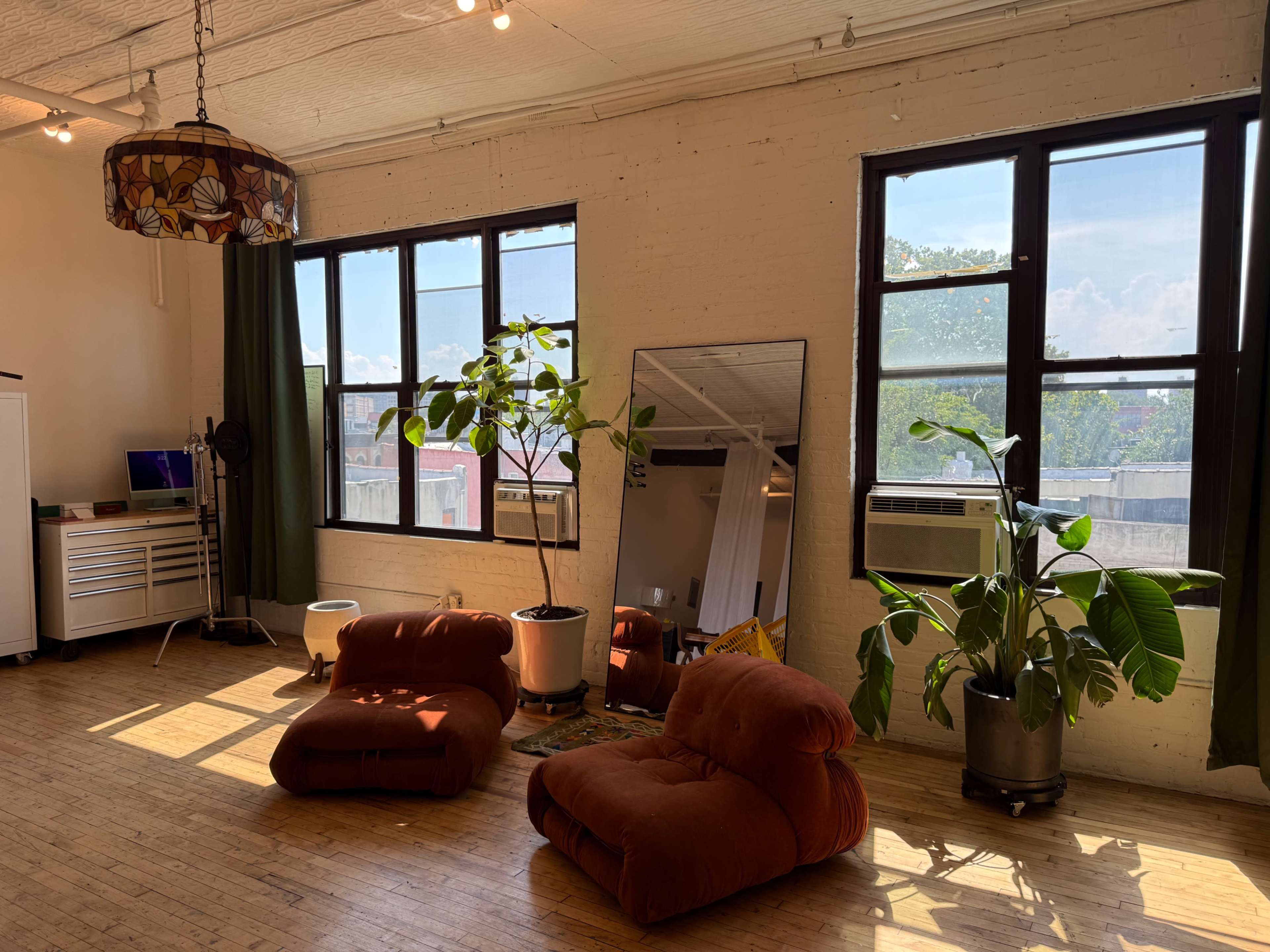 The image shows a bright, spacious room with large windows, a mirror, two plush chairs, and potted plants.