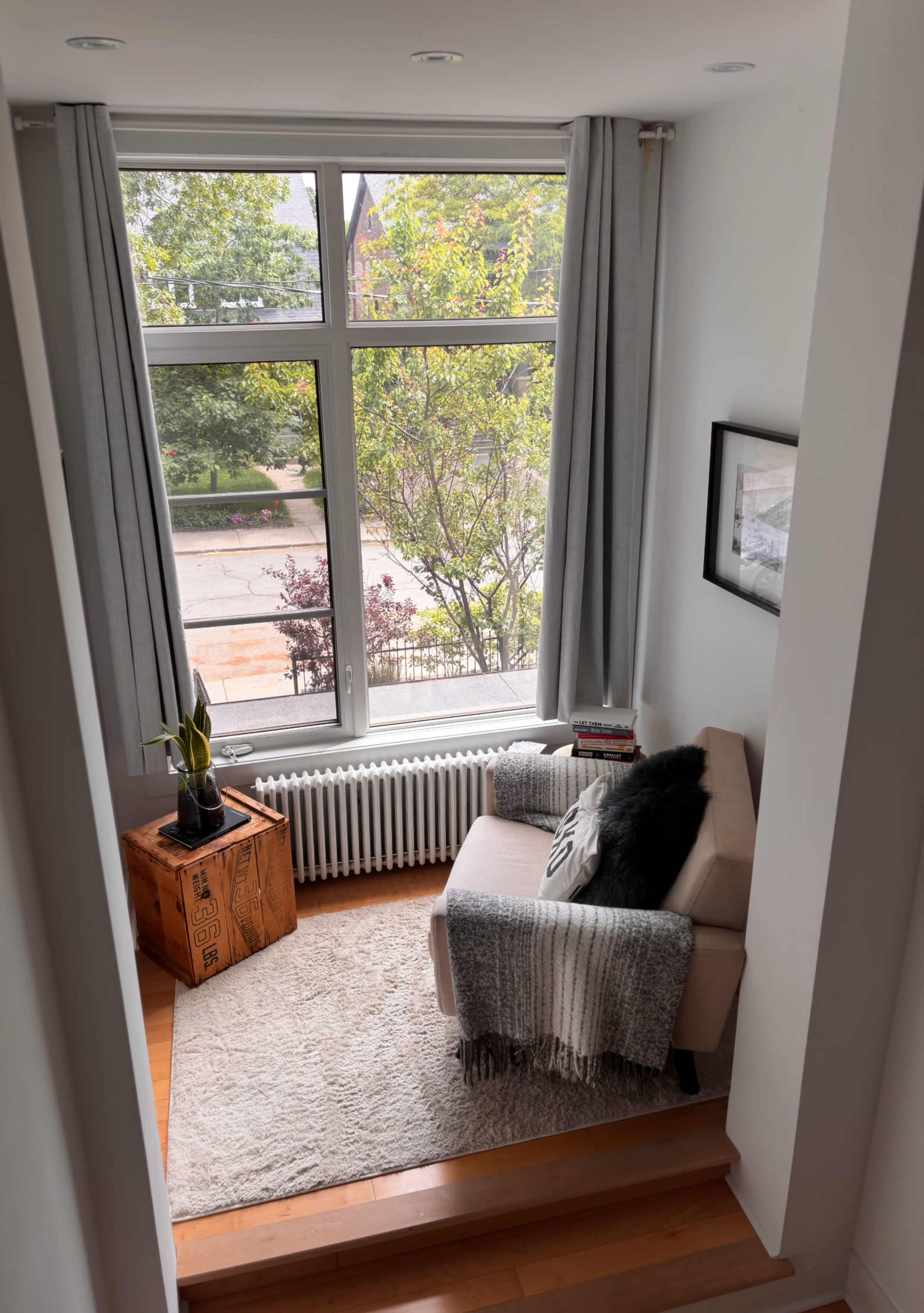 A cozy corner of a home features an armchair, a small table, and a large window overlooking a treed street.