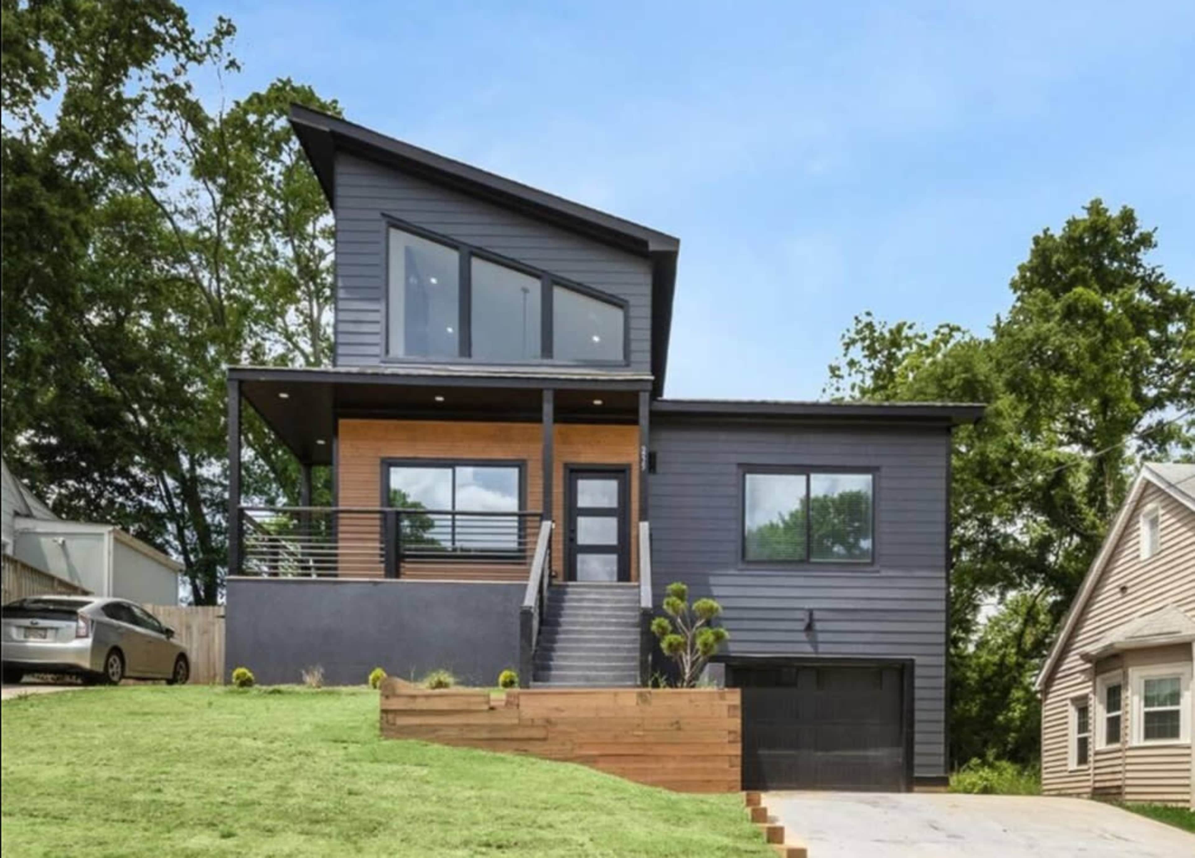 The image shows a modern two-story house with a dark exterior, large windows, and a front staircase leading to a deck.