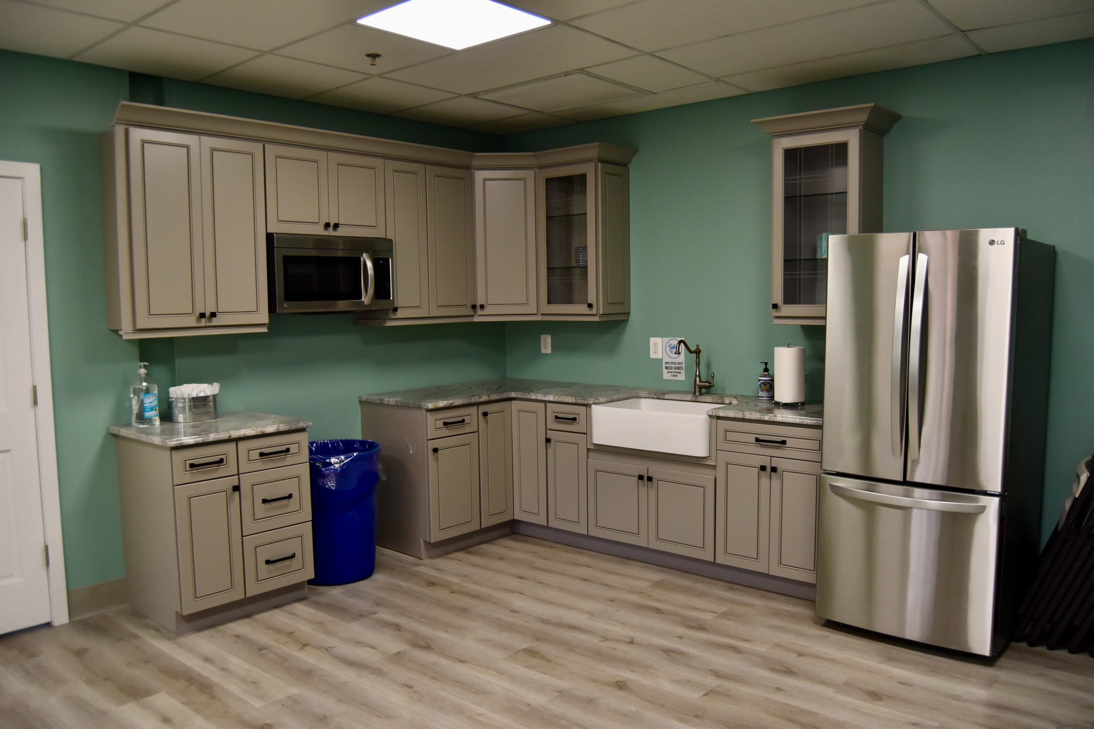 A kitchen featuring gray cabinetry, a stainless steel refrigerator, a large white sink, and light wood flooring against a teal wall.