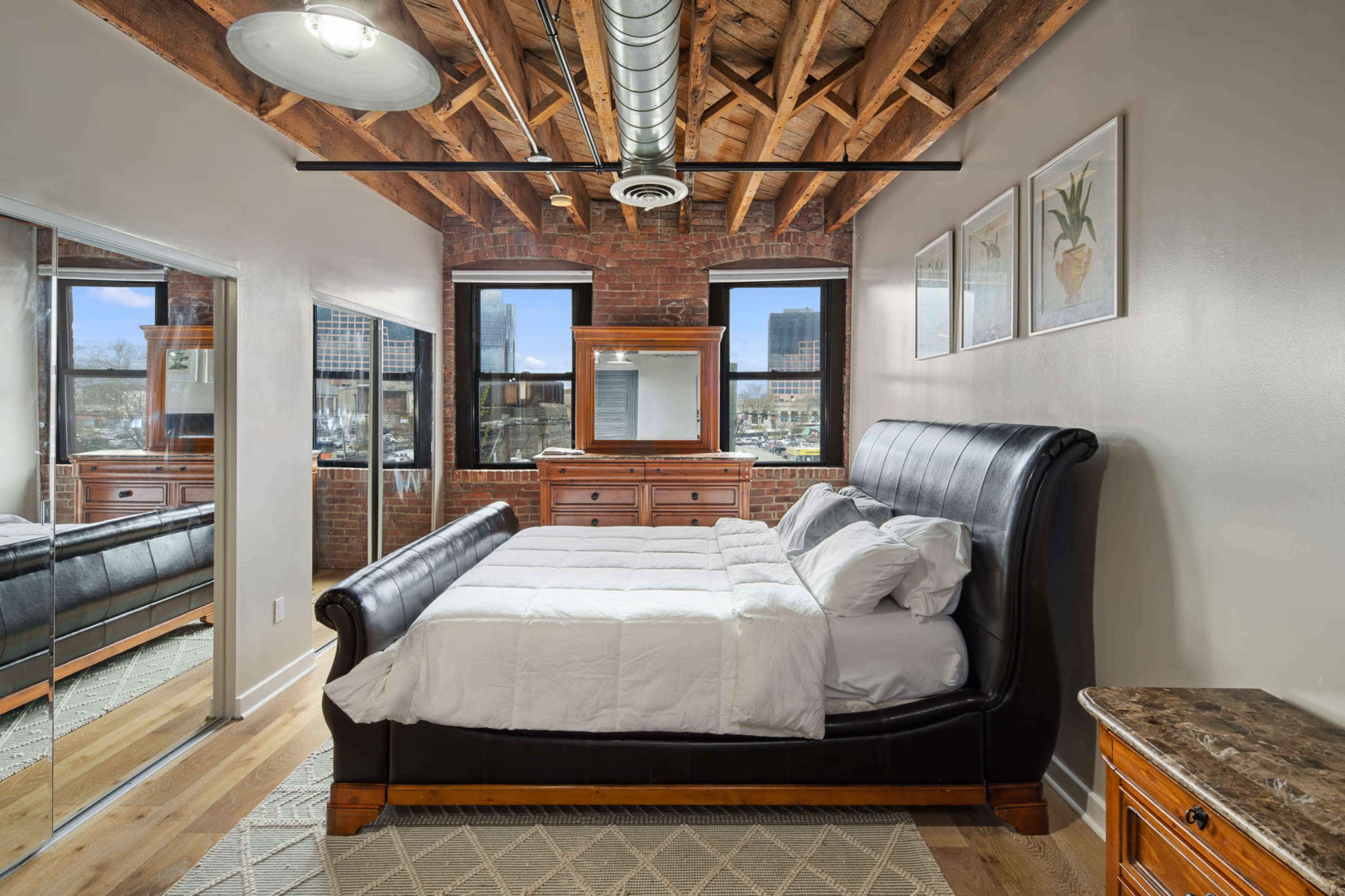 The bedroom features a black leather sleigh bed, wooden furniture, exposed brick walls, and large windows with city views.
