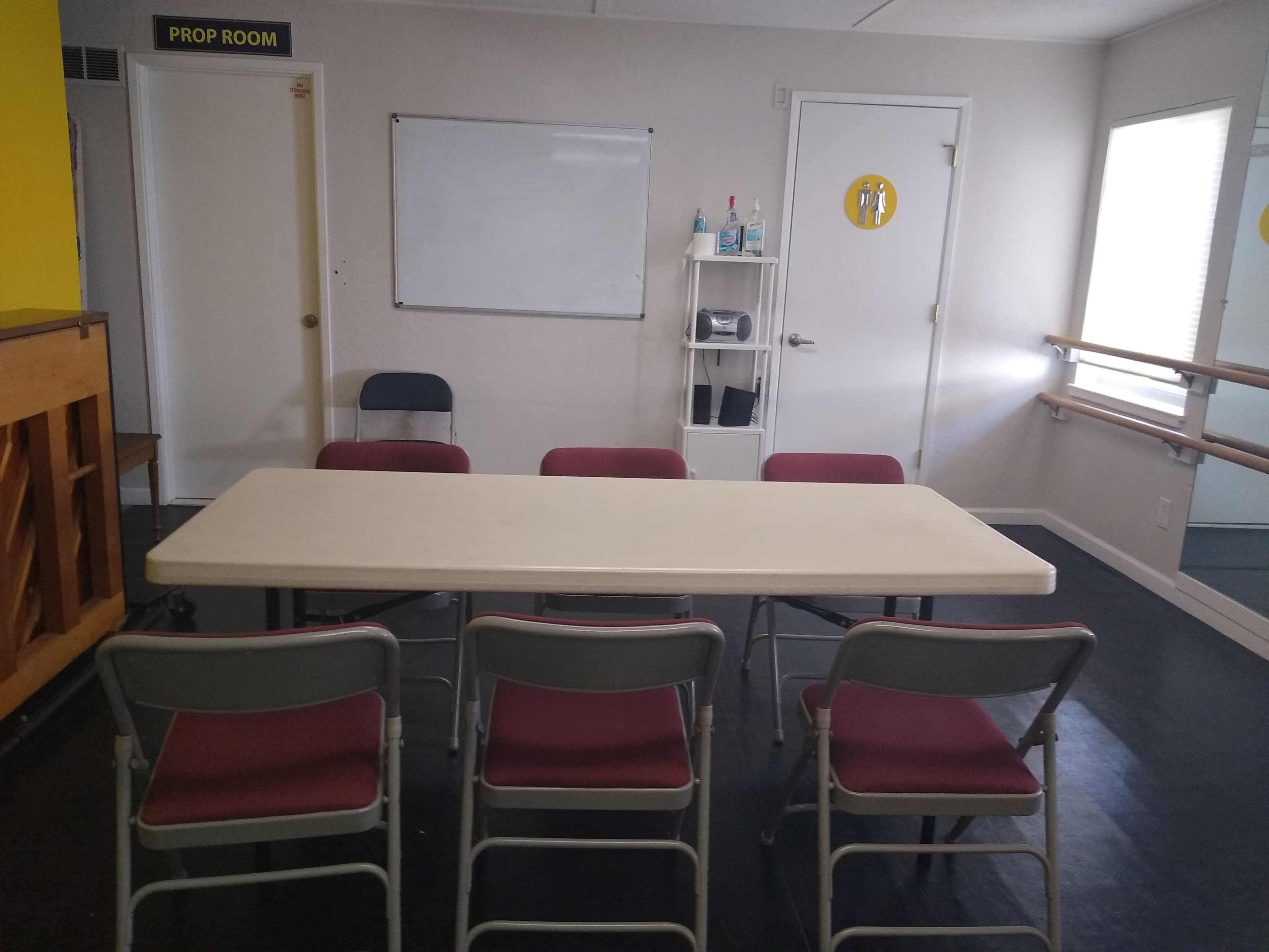 A rectangular table with six chairs is positioned in a room that includes a whiteboard and a shelf with items ready for use.