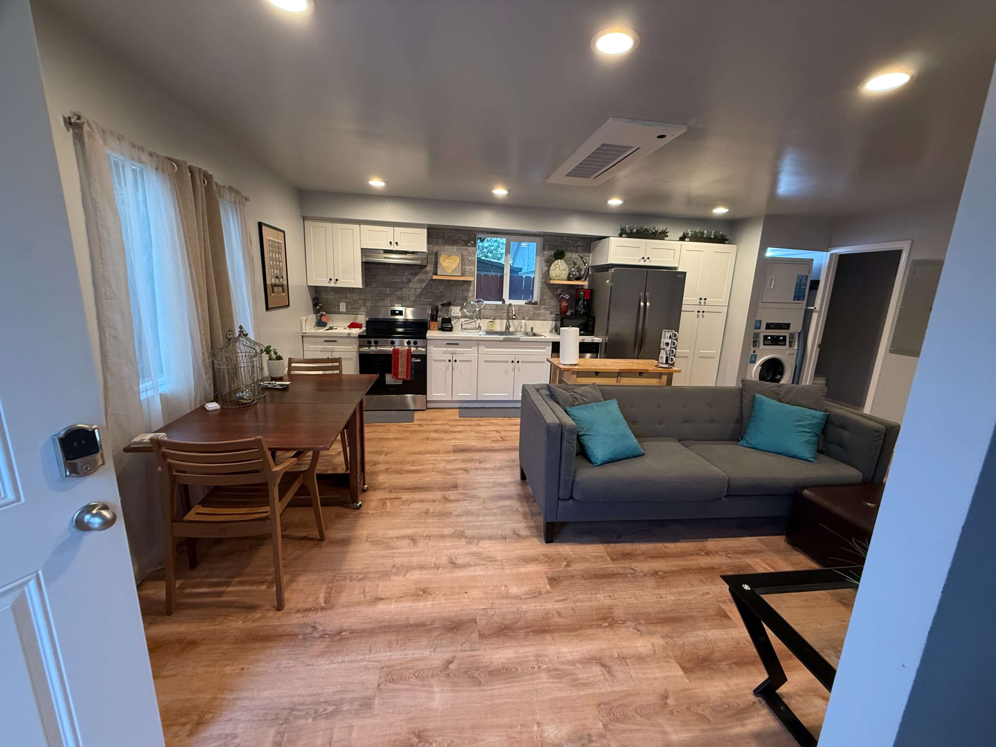 The image shows a modern kitchen and living area with a gray sofa, a dining table, and appliances.
